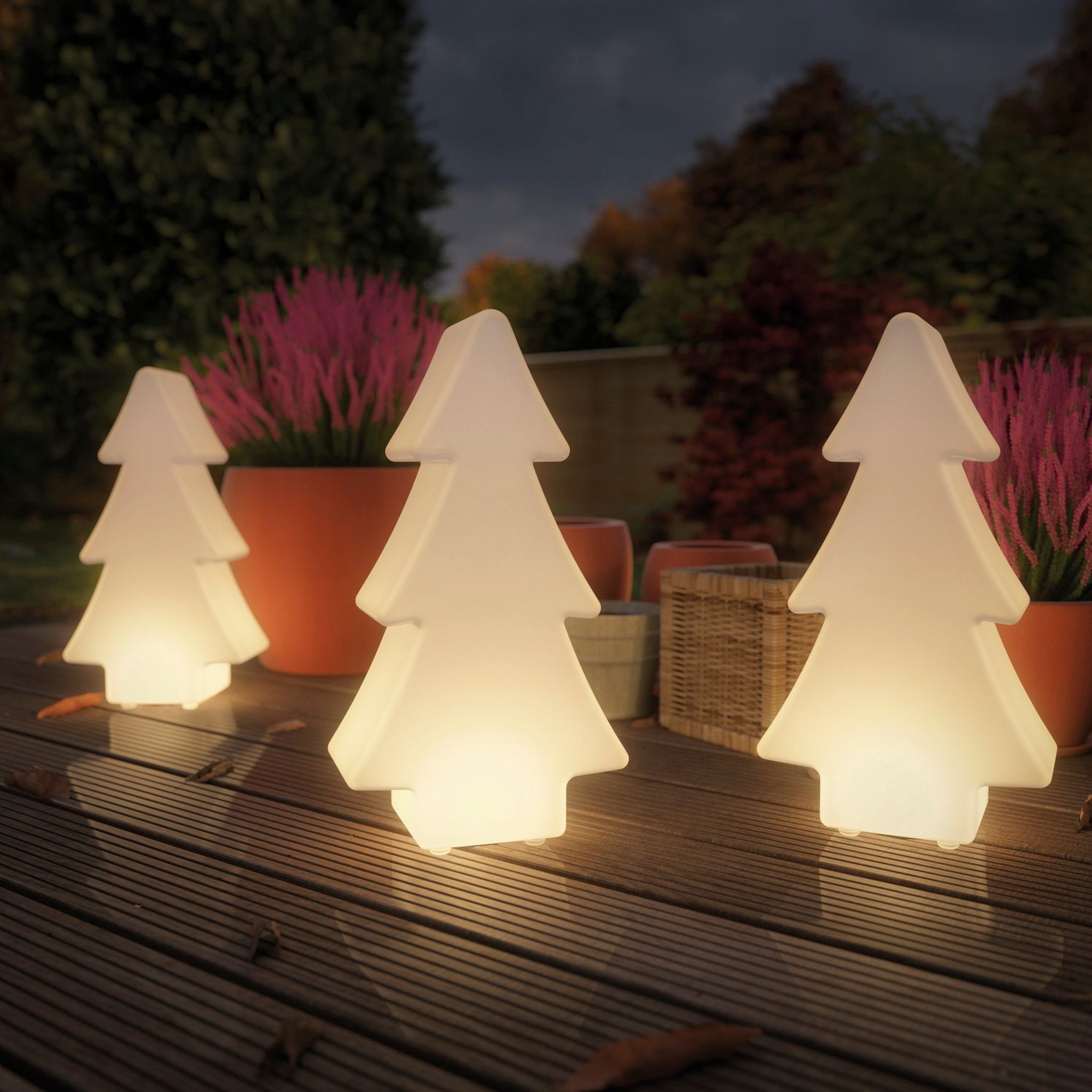 Three Paulmann Plug & Shine Outdoor Christmas Tree 2.8W LED Floor Lamps with Ground Spikes in White, resembling illuminated fir trees, adorn a wooden deck surrounded by potted plants, enhancing the charm of this serene outdoor space. Thanks to Plug & Shine's convenience, they effortlessly illuminate the garden in the background.
