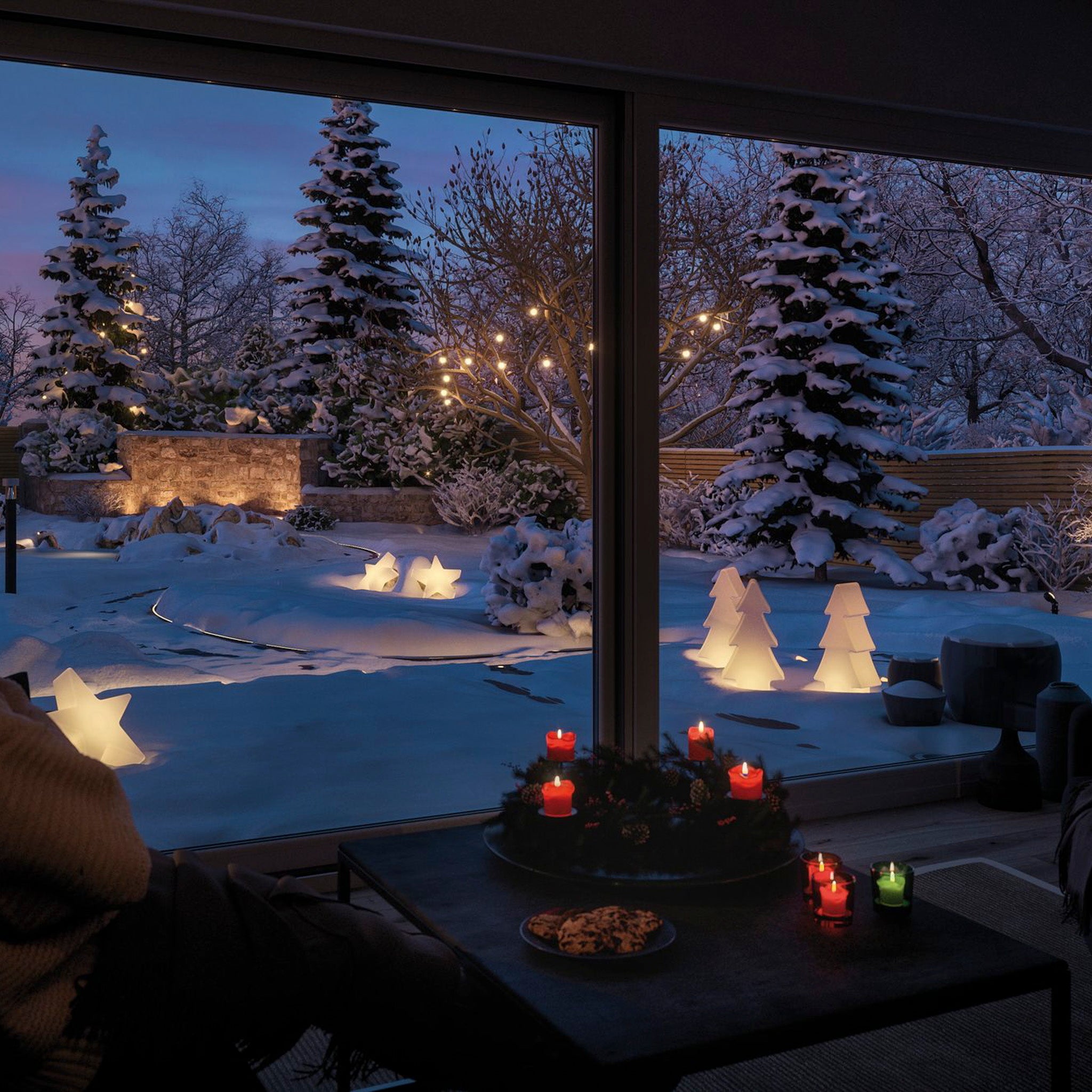 A cozy room with candles overlooks a snowy garden, where an illuminated fir tree and glowing star decorations create a magical atmosphere at dusk. Experience the enchantment as the Paulmann - Plug & Shine Outdoor Christmas Tree 2.8W LED Floor Lamp with Ground Spike in White transforms outdoor spaces into winter wonderlands.