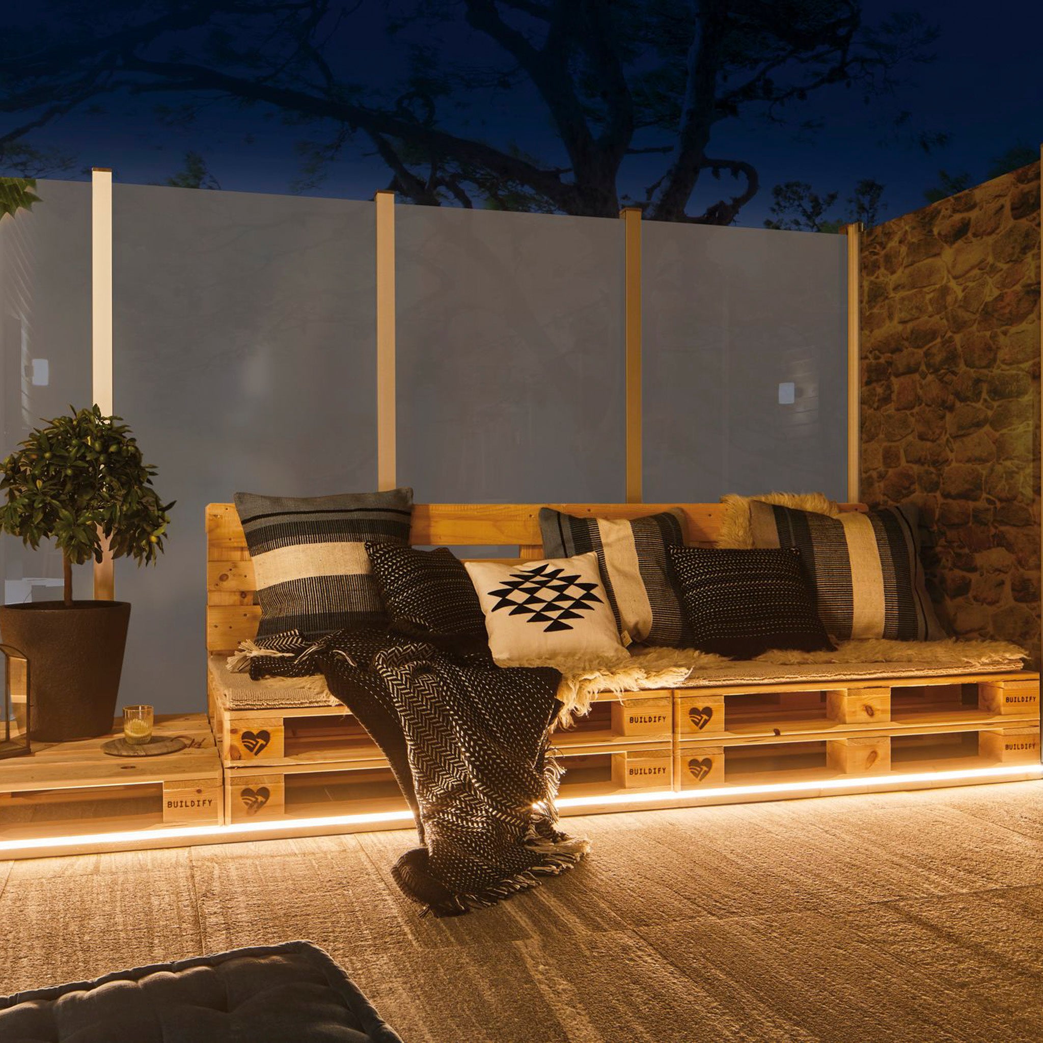 Create a cozy outdoor seating area using pallets complemented by cushions and blankets, enhanced by a weather-resistant setting with potted plants against a stone wall. The Paulmann - Plug & Shine Outdoor 31W LED Light Strip in Warm White provides soft, inviting lighting for the perfect ambiance.