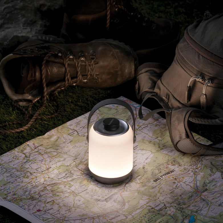 A portable lighting setup: the Paulmann Outdoor Clutch 6W LED Mobile Table Lamp in Grey casts a pleasant light over a map, with hiking boots and a backpack nearby, all set on the grass at dusk.