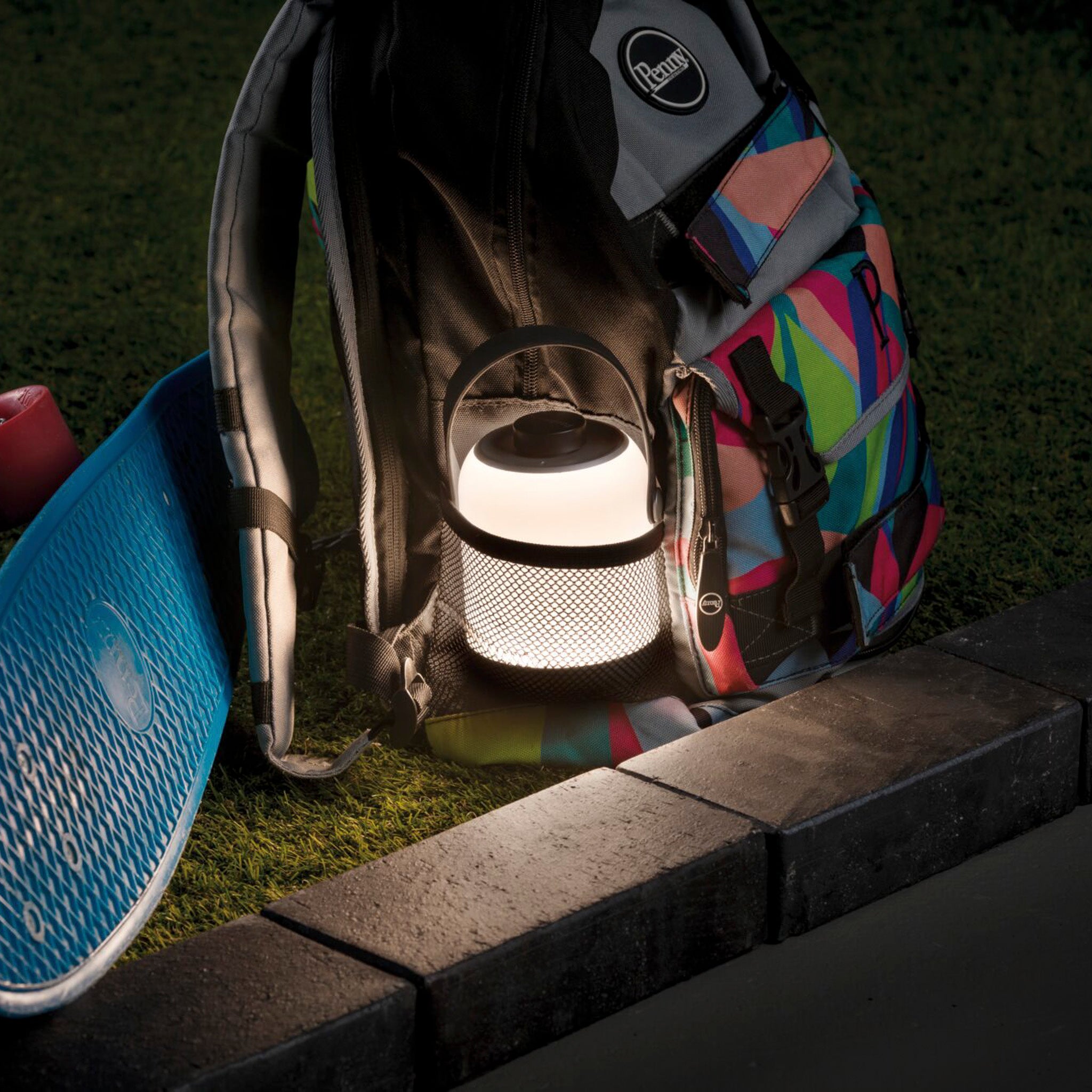A Paulmann Outdoor Clutch 6W LED Mobile Table Lamp in Grey casts a pleasant light inside a colorful backpack next to a skateboard on the grassy surface.