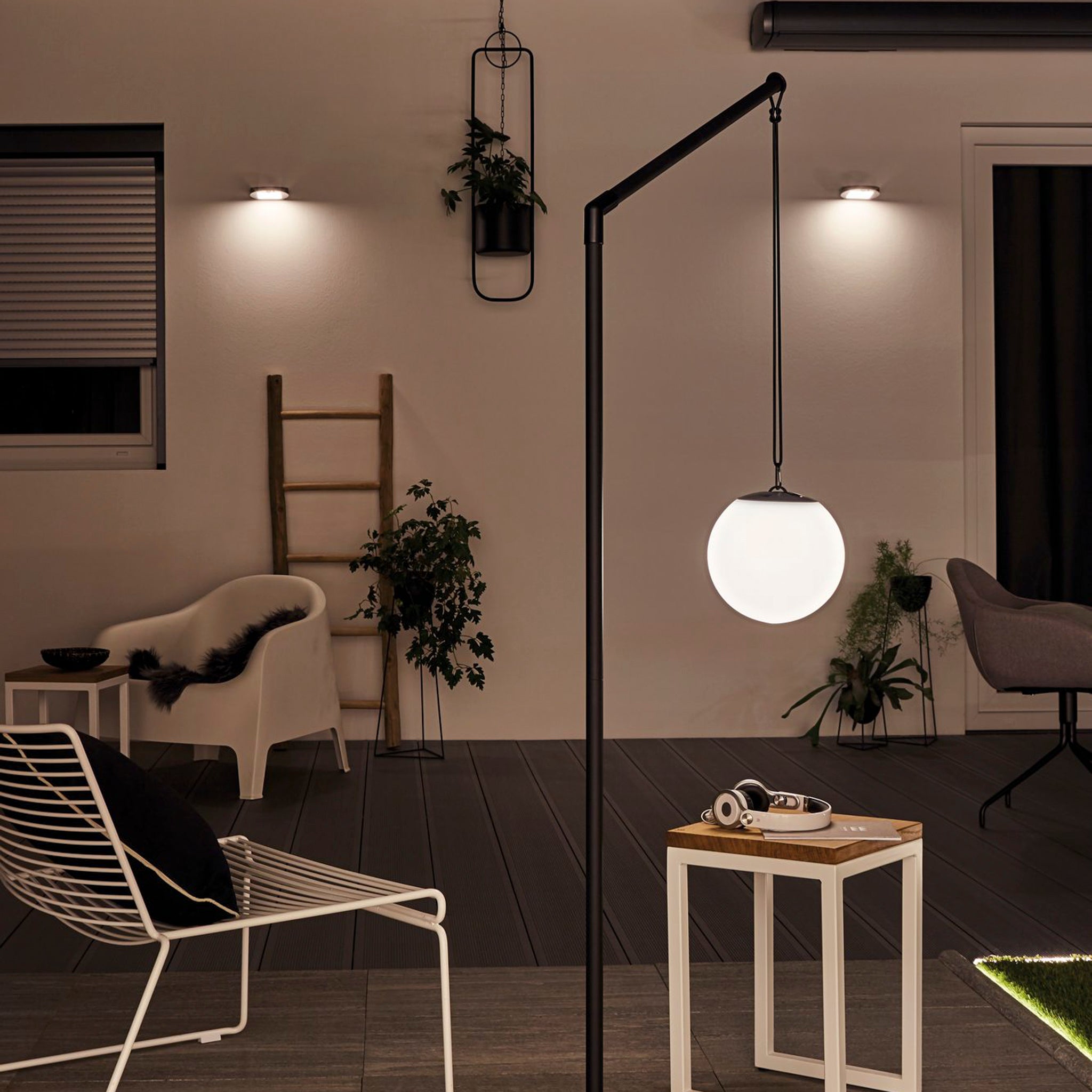 A contemporary patio featuring chairs, the Paulmann Ryse 1.2W LED solar wall light with motion detection in white casting a gentle glow, small plants, and a wooden table with headphones beneath stylish exterior walls.