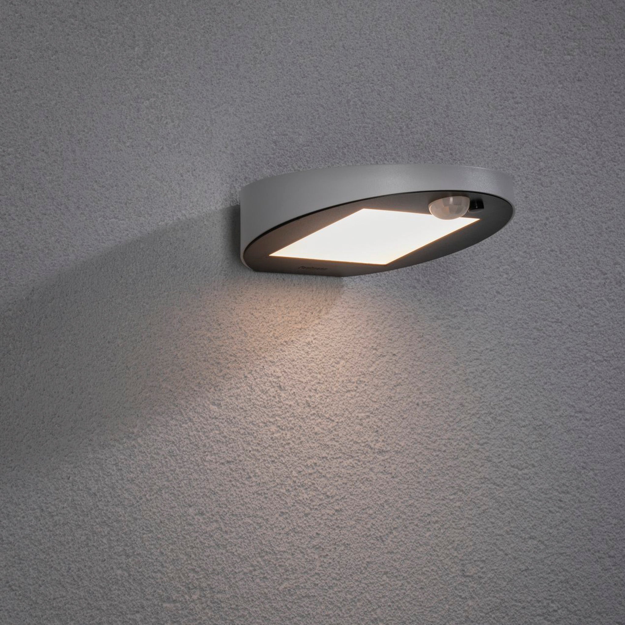 The Paulmann Outdoor Ryse 1.2W LED Solar Wall Light in white, featuring motion detection, subtly illuminates against a textured gray wall.