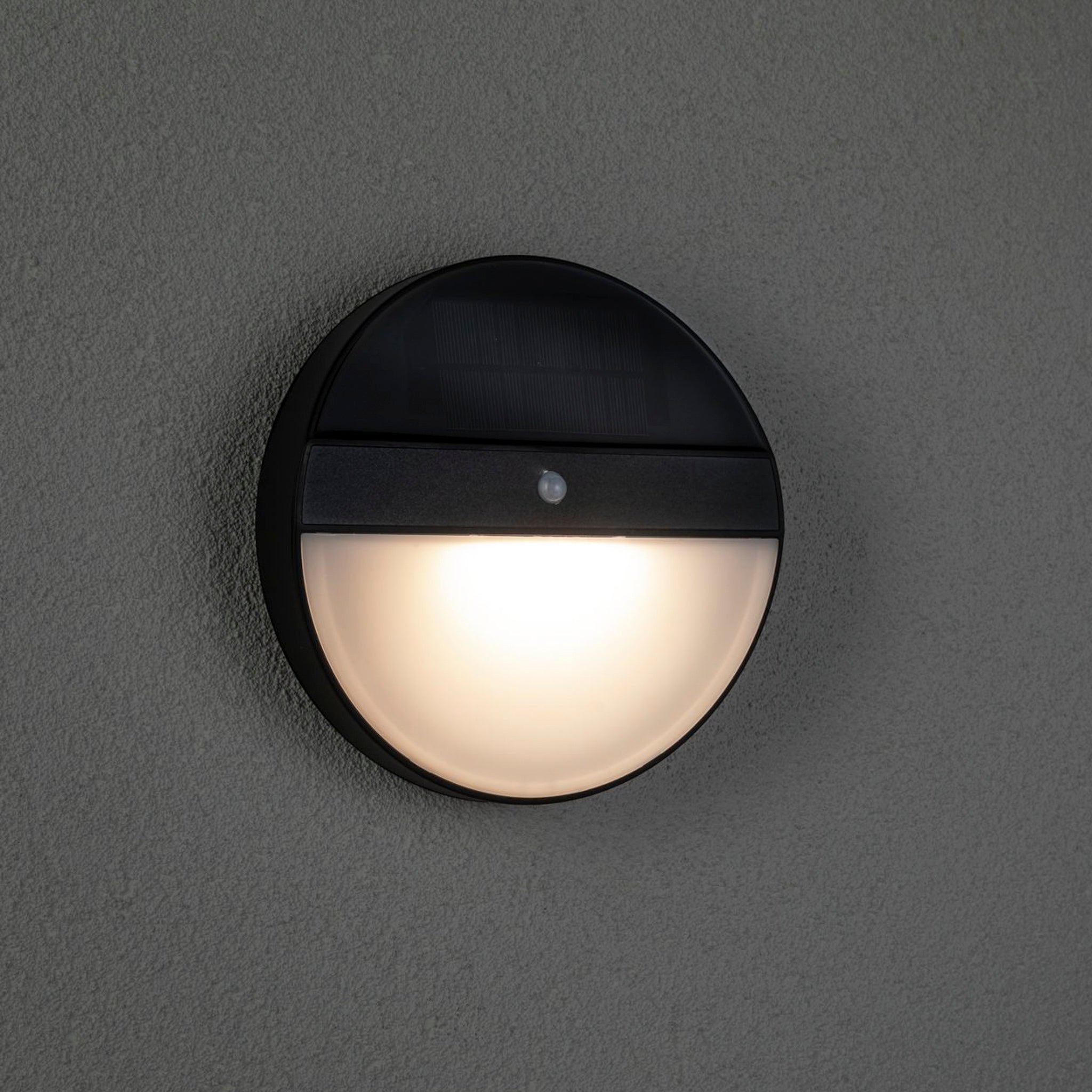 The Paulmann Outdoor Elios 1W LED Solar Wall Light with Motion Detection in Anthracite emits a warm glow on a textured gray exterior wall.