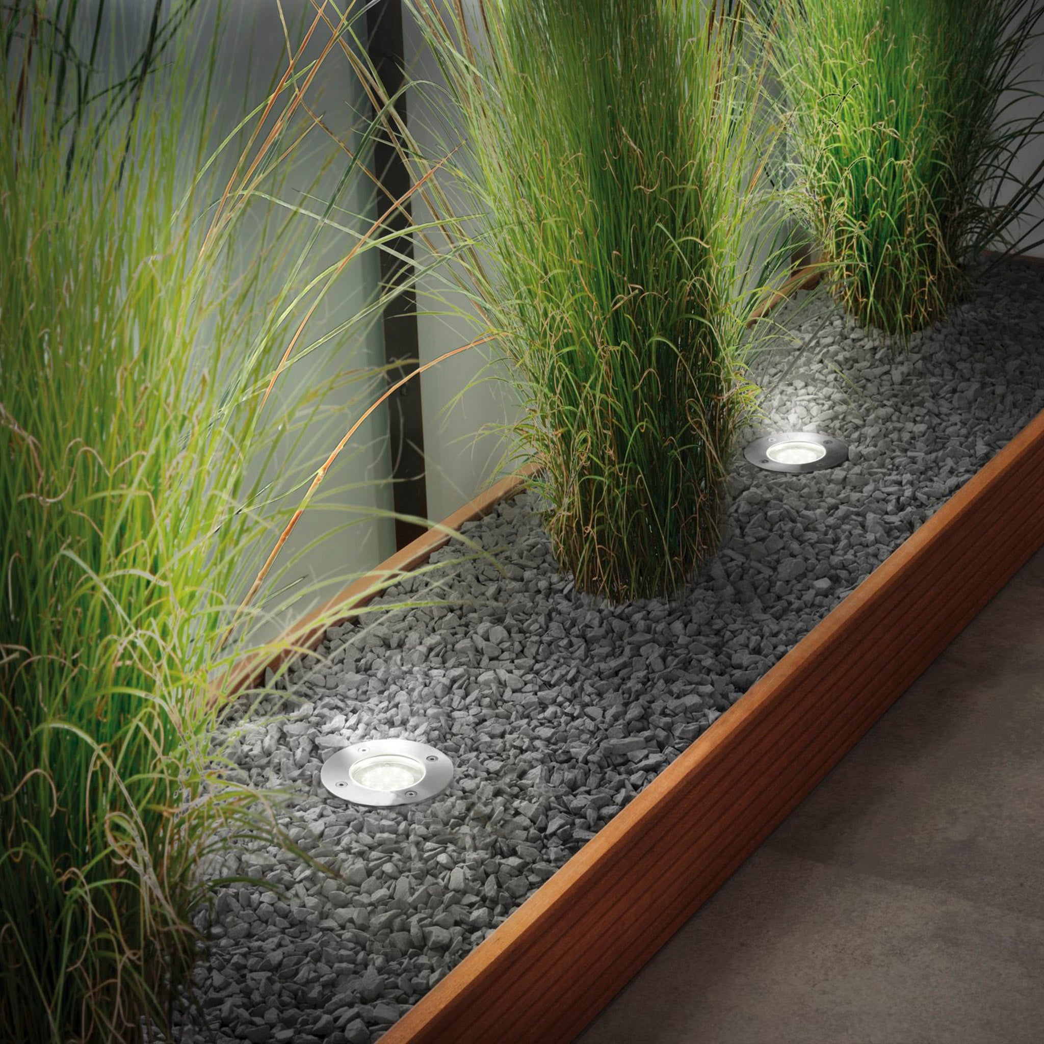 An outdoor planter with tall grass is illuminated by the Paulmann - Plug & Shine Outdoor 3.6W RGBW Floor Mini Individual Spotlight in silver, placed on gray pebbles and bordered by wood.