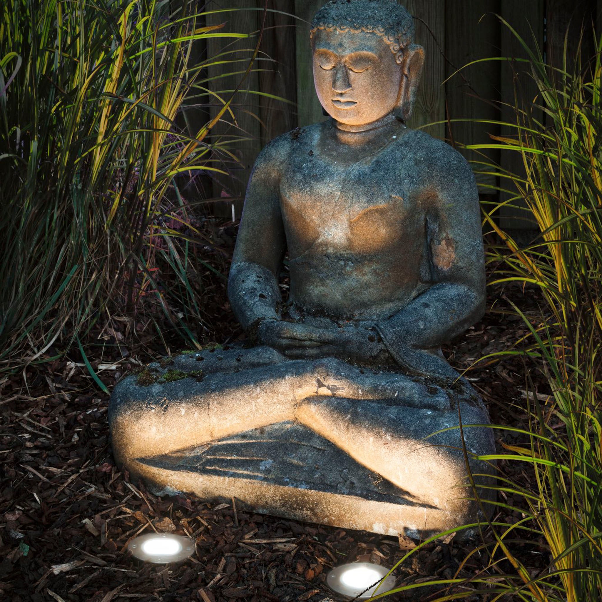A weathered stone Buddha statue sits cross-legged in the garden, surrounded by grass and gently illuminated at night by a Paulmann - Plug & Shine Outdoor 3.6W RGBW Floor Mini Individual Spotlight in Silver.