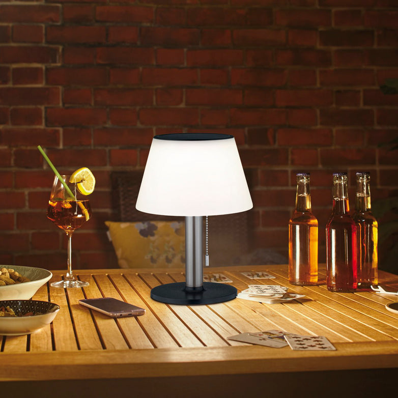 The Paulmann Outdoor Lillesol 0.8W LED Solar Table Lamp with a 3-step dimming feature in stainless steel gently casts light over a wooden table adorned with drinks, playing cards, and snacks in a softly lit room, evoking the warmth of cozy garden lighting.