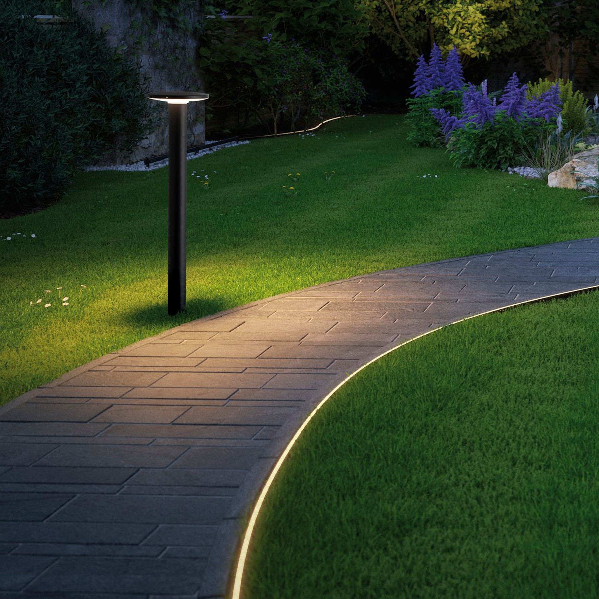 A curved stone pathway with Paulmann Plug & Shine 6.1W LED Bollard Lights in anthracite, offering a warm white glow and 360-degree beam angle, is bordered by a manicured lawn thriving amidst lush greenery and purple flowers, creating a gentle illumination at dusk.
