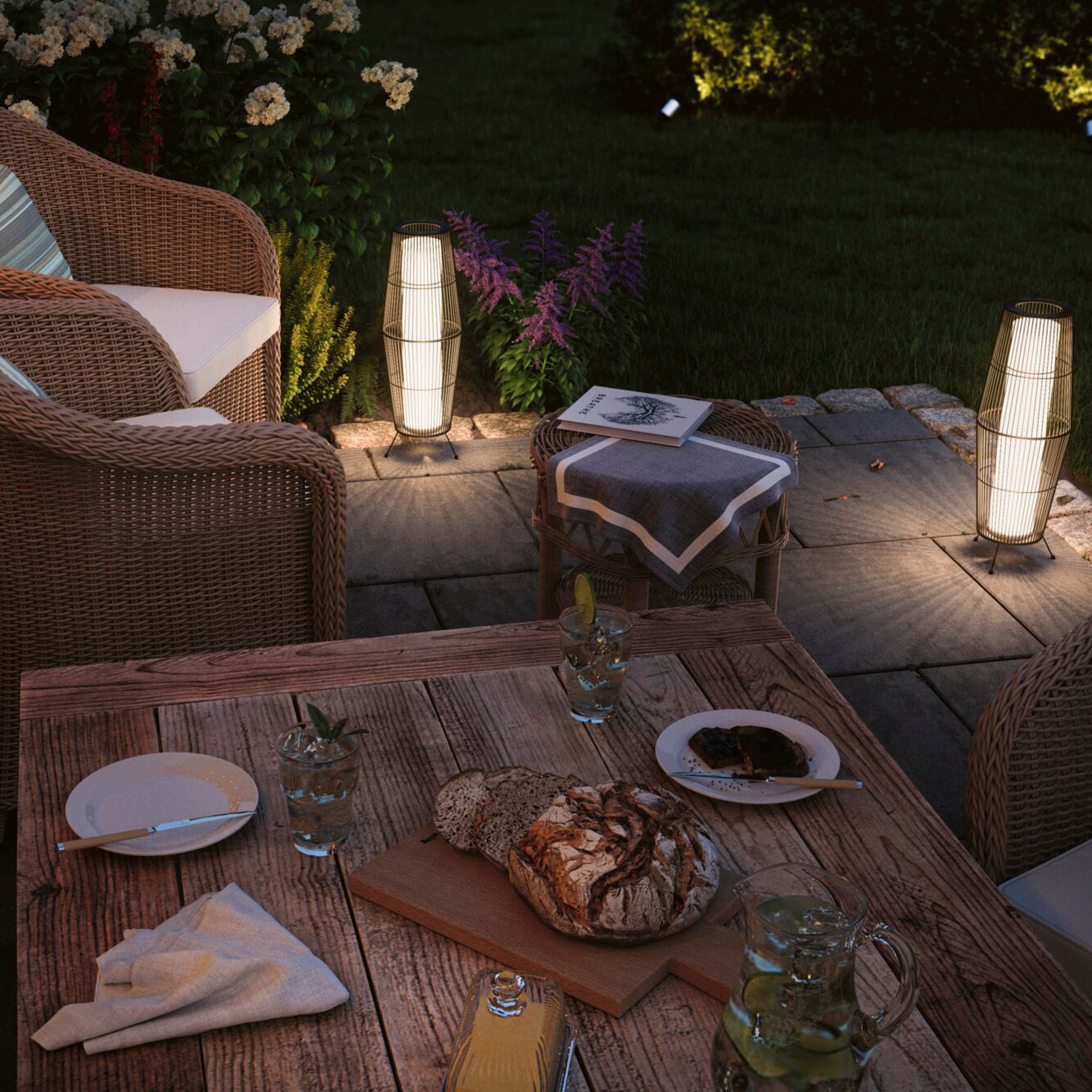 A cozy outdoor patio featuring wicker furniture, bread on the table, drinks, and the warm illumination of the Paulmann Plug & Shine Outdoor Basket 7.8W LED Floor Light in Brown lighting up the night.