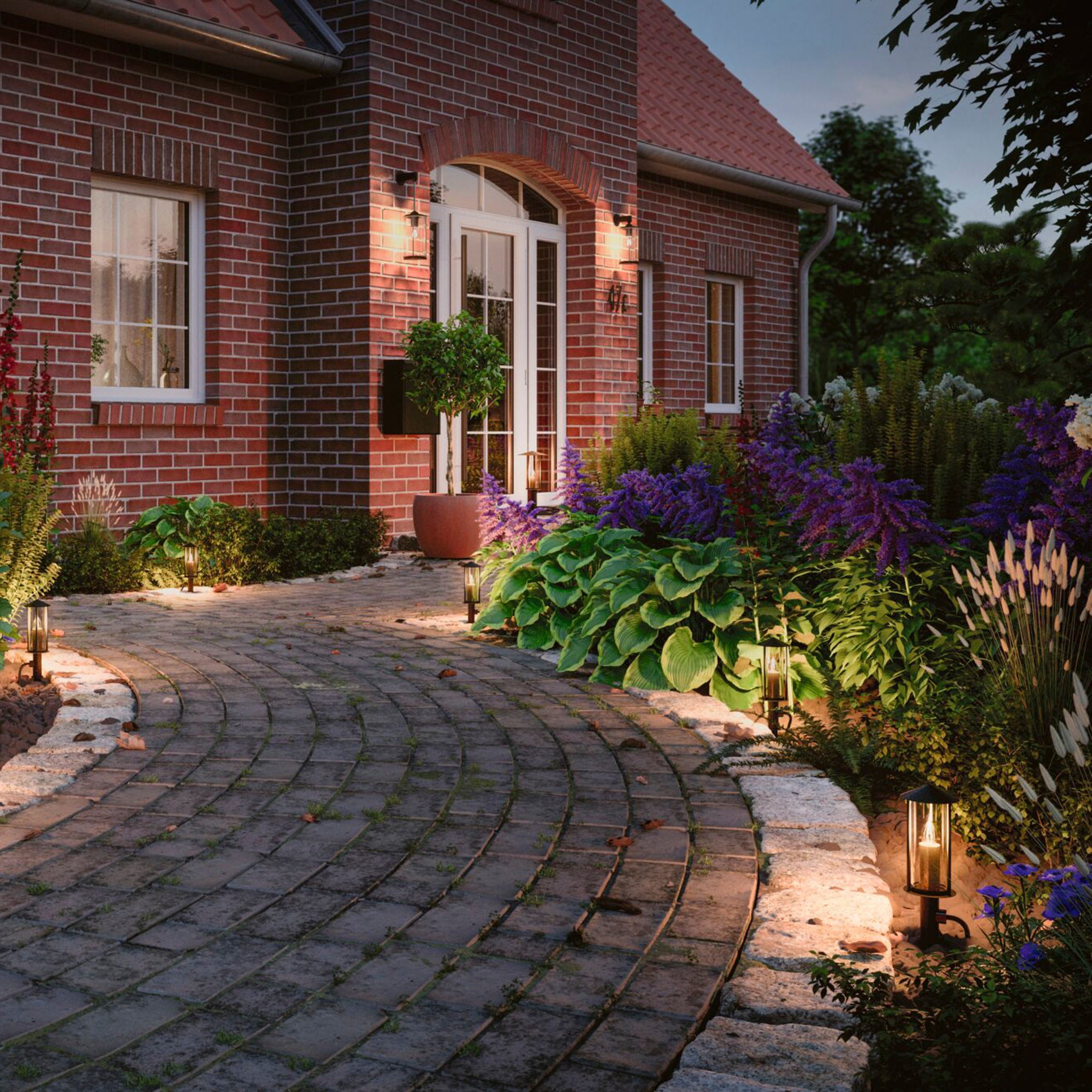 A red-brick house features a charming garden path illuminated at dusk by the subtle glow of 50cm bollard lights from the Paulmann Plug & Shine Outdoor Classic series, surrounded by lush green plants and vibrant purple flowers.