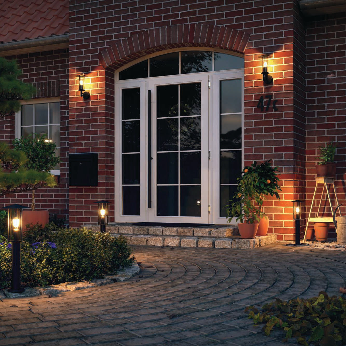 The entrance of the brick house with white doors and potted plants is beautifully complemented by Paulmann's Outdoor Classic 15W LED Bollard Light in Dark Grey, providing warm IP44 protection lighting along the cobblestone pathway at dusk to enhance its modern appearance.