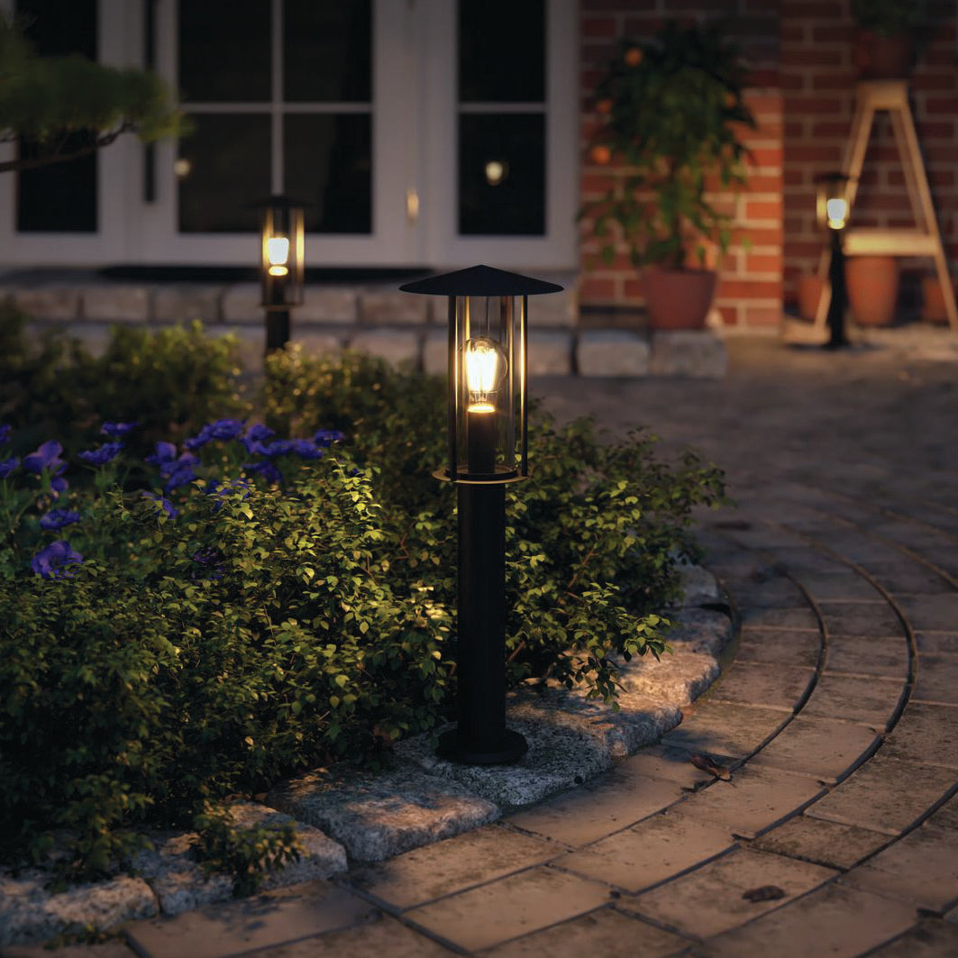 The garden pathway at night, adorned with lit lanterns and lush greenery, exudes a modern look. The way is illuminated by Paulmann's Outdoor Classic 15W LED Bollard Light in Dark Grey, featuring IP44 protection, while a brick building quietly stands in the background.