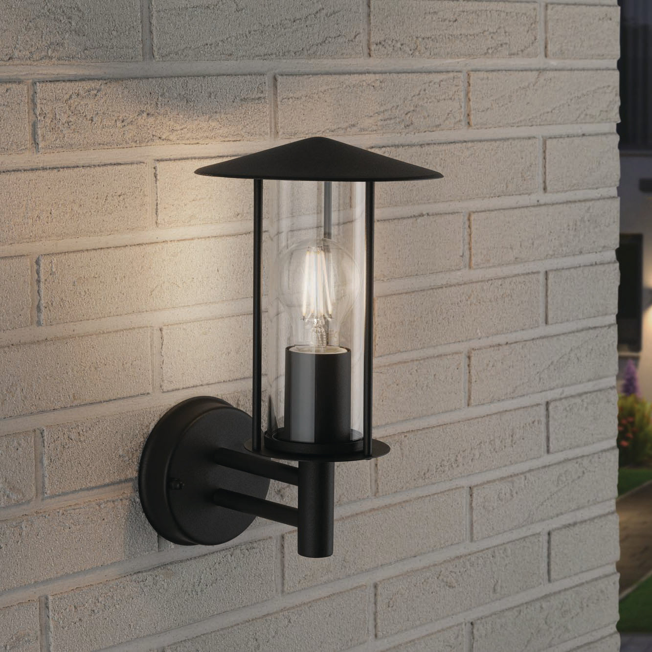 Paulmann's Outdoor Classic 15W LED Wall Light in Dark Grey is mounted on a brick wall, offering IP44 protection and emitting a warm glow at night.