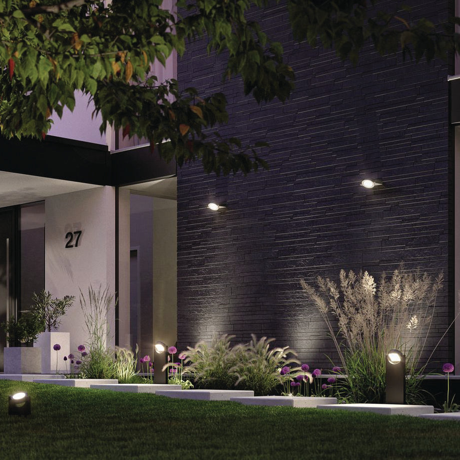 The contemporary building exterior is enhanced by the Paulmann - Outdoor Swivea 8W LED Wall Light in Anthracite, elegantly illuminating the area and showcasing the plants and house number 27. Nestled among trees at night, these cardanic swiveling fixtures contribute a refined touch to its architectural sophistication.