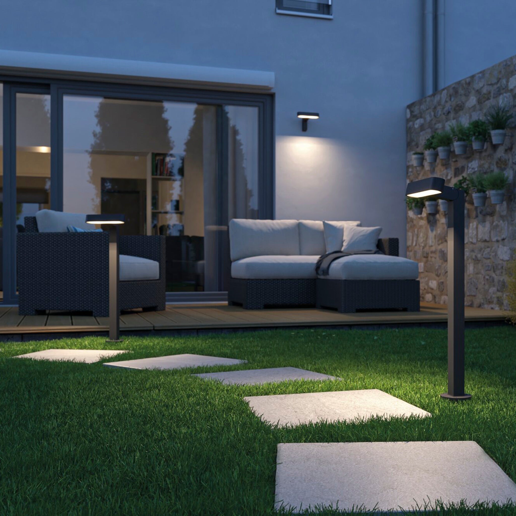 A modern patio with wicker sofas and a stone path is beautifully lit by Paulmann's Plug & Shine Outdoor Ito 6W LED Horizontal Bollard in Anthracite. These lights subtly enhance the ambiance against a house with large windows, creating an inviting evening retreat.