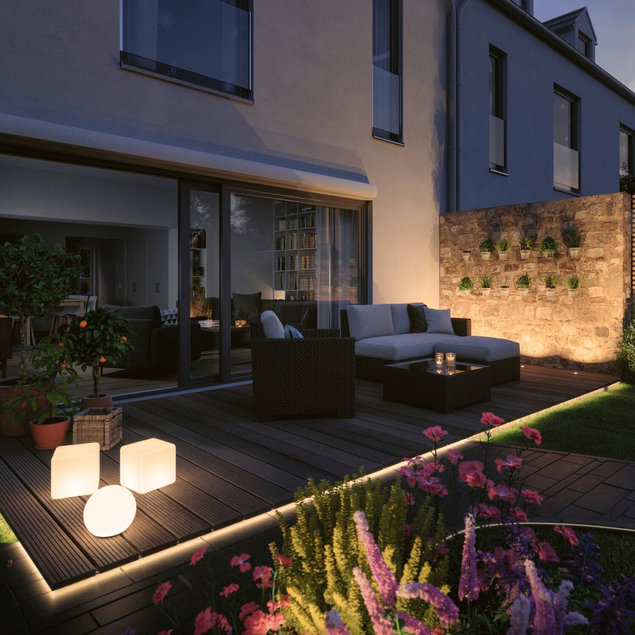 A contemporary patio featuring comfortable seating, illuminated with Paulmann's Plug & Shine Outdoor 48W LED 10m Smooth Strip in RGBW that integrates with Smart Home Zigbee 3.0 for a vibrant glow, is accompanied by cube lights and a vertical garden on a stone wall. The ambiance is enhanced with subtle garden illumination and surrounded by an array of lively flowers.