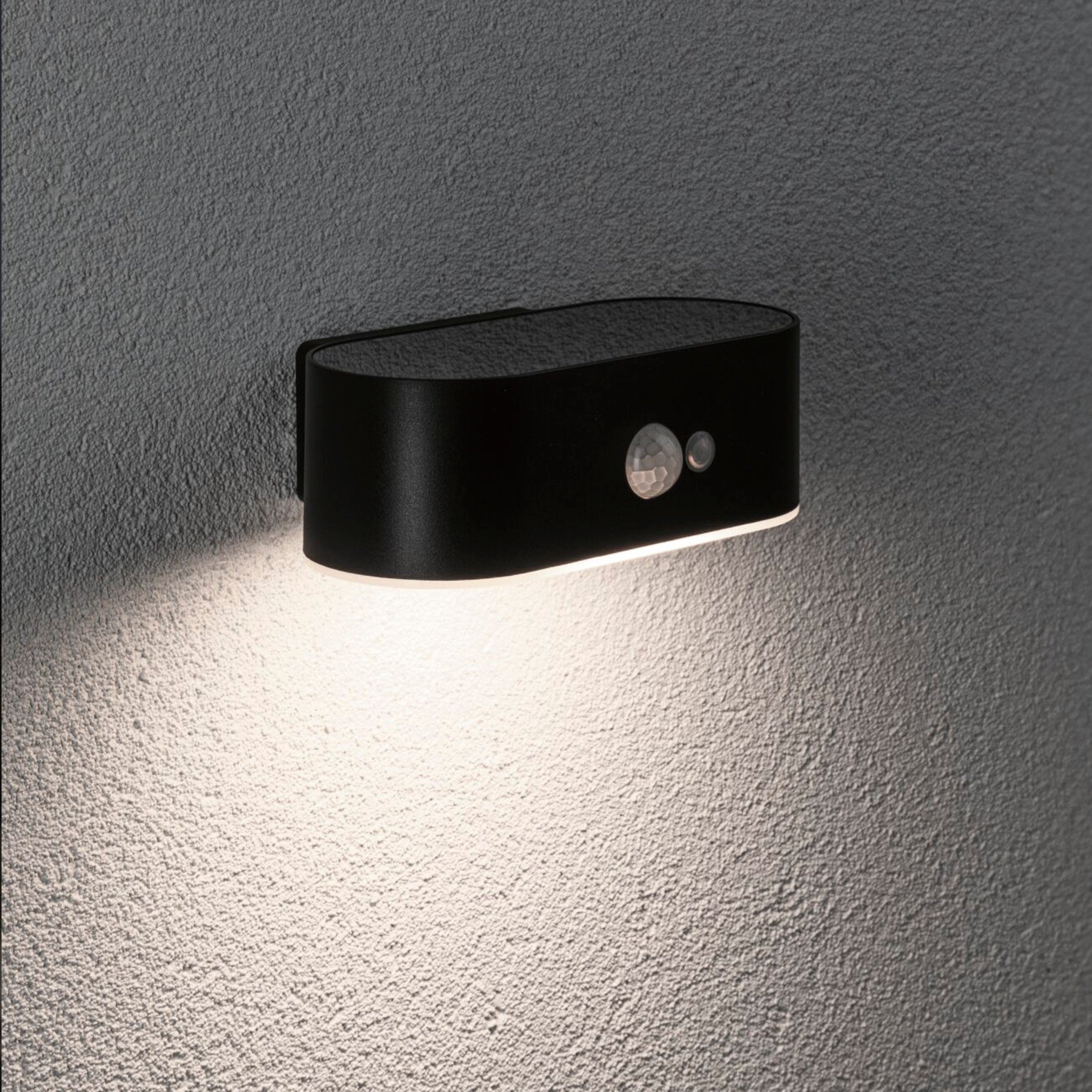The black Paulmann Outdoor Adya 2.5W LED Solar Wall Light, featuring energy-saving technology and motion detection, casts light downward on a gray surface.