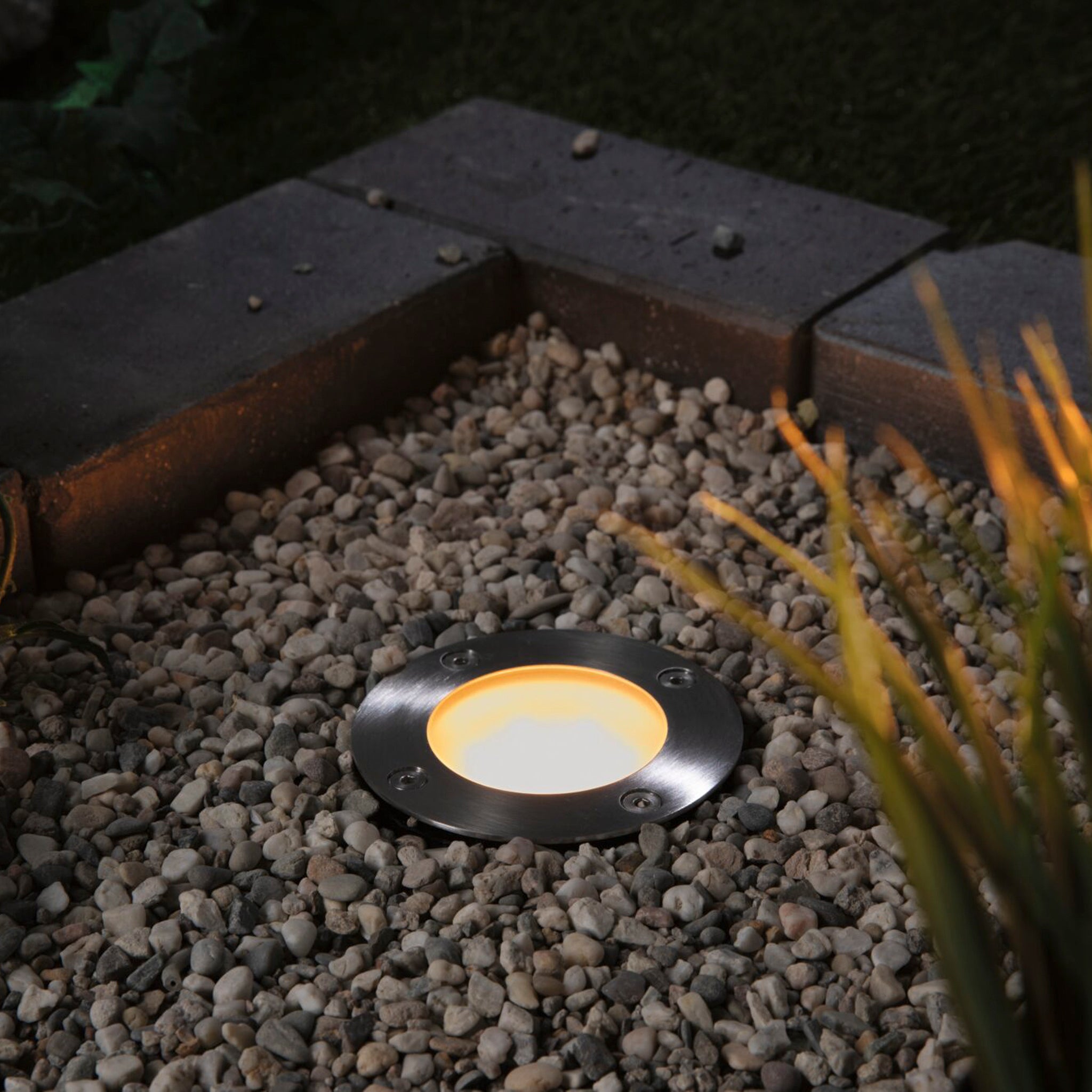 The Paulmann - Plug & Shine Outdoor 5.2W LED Recessed Spotlight in Black, compatible with Smart Home Zigbee 3.0, is installed at ground level among stones, illuminating nearby grass and plants during dusk.