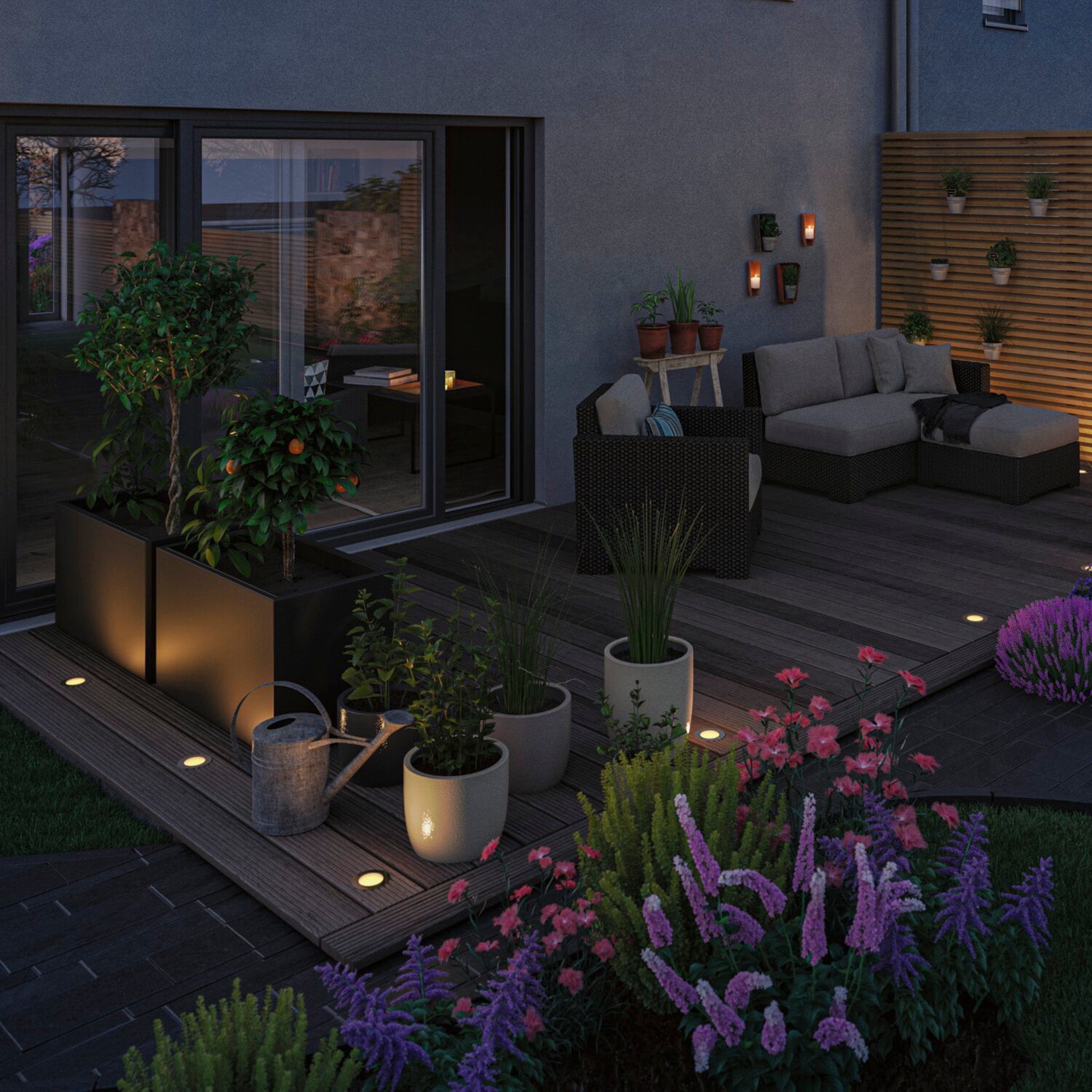 Transform your patio into a cozy retreat at dusk with potted plants and a seating area enhanced by ambient lighting and blooming flowers, all brightened by the Paulmann - Plug & Shine Outdoor 5.2W LED Recessed Spotlight in black. Elevate the atmosphere with its Smart Home Zigbee 3.0 technology for seamless illumination control.
