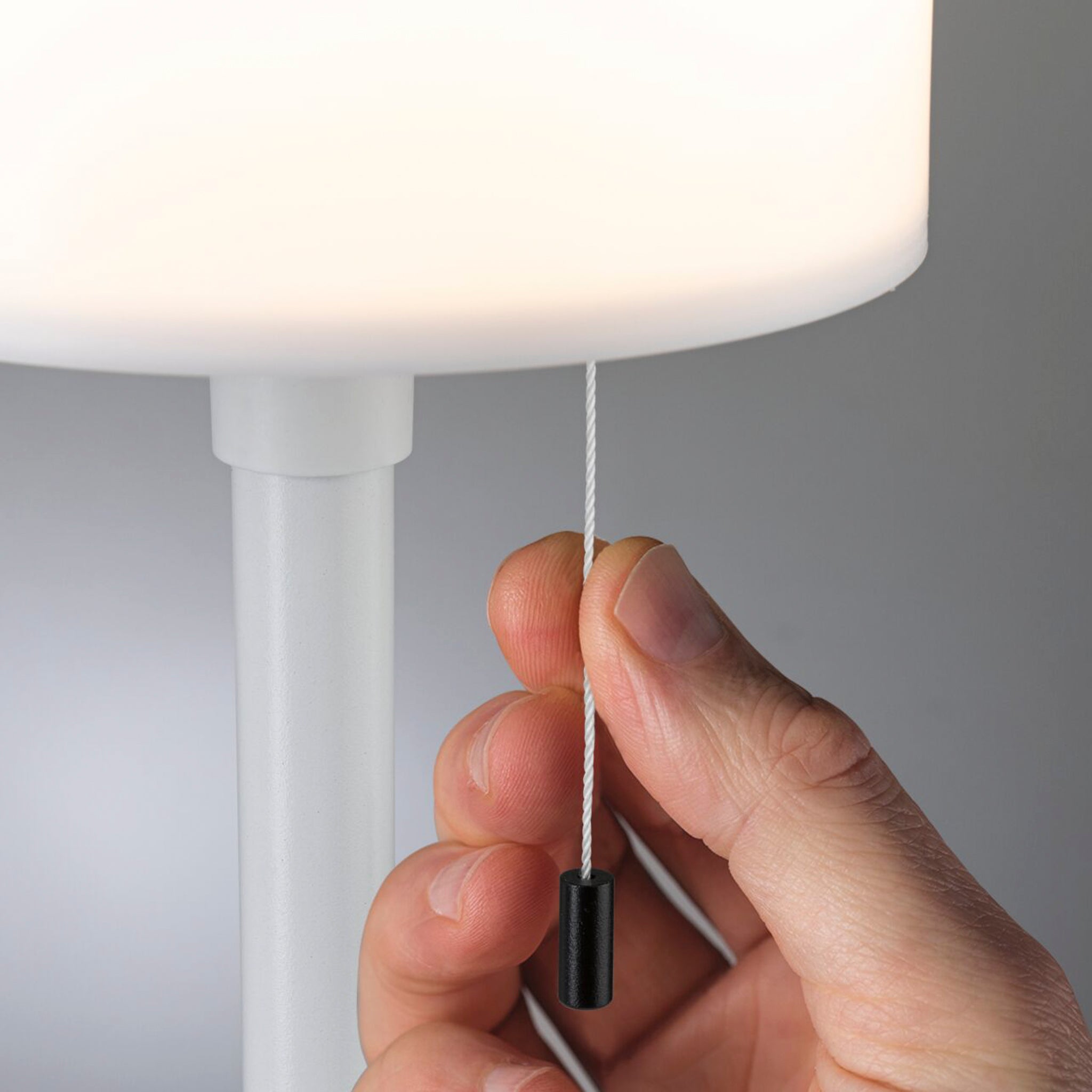 A close-up of a hand pulling a cord to turn on the sleek, white *Paulmann - Bartja 1.6W LED Solar Table Lamp with 3-Step Dim*, featuring IP44 protection and ideal for any environment.
