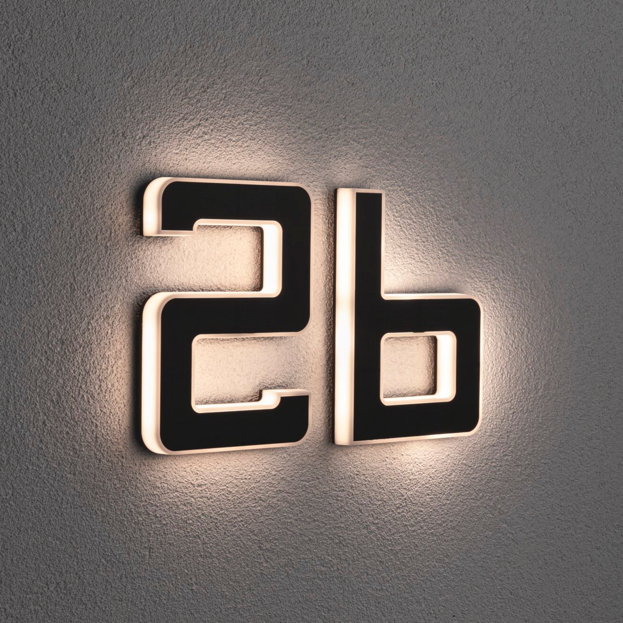 The Paulmann Outdoor Solar House Letter "B" LED 0.2W luminaire with a replaceable battery in black softly glows on a textured wall, creating a gentle backlight effect.
