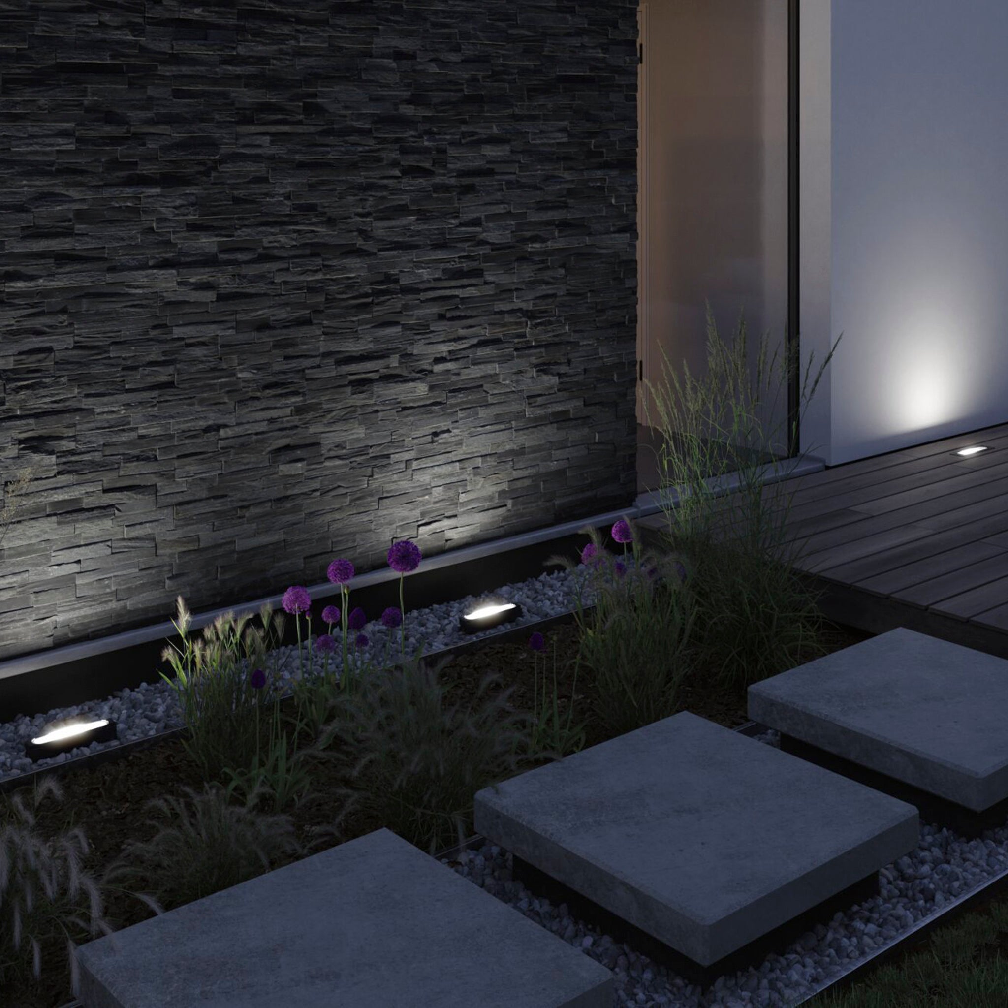 A contemporary garden path featuring concrete slabs, vibrant purple flowers, and a stone wall illuminated by warm white lighting at night is enhanced with the discreet integration of Paulmann - Plug & Shine Outdoor Flotas 6.1W LED Wallwashers in Anthracite. These recessed luminaires enrich the design and create an inviting ambiance ideal for relaxing evenings.
