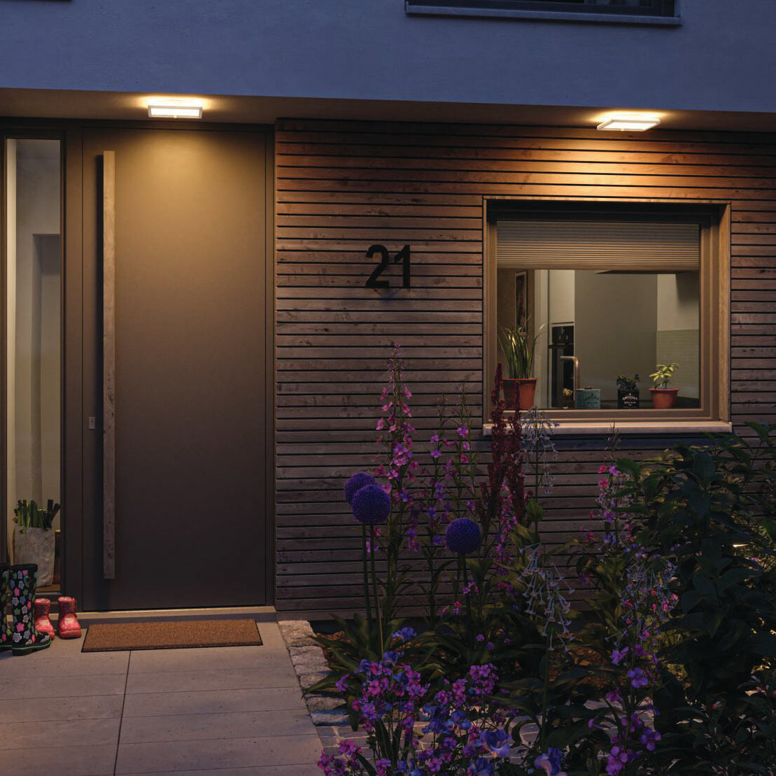 A welcoming house entrance featuring a wooden facade is beautifully lit by the Paulmann Outdoor Lamina Insect Friendly 16W LED Backlit Square Tunable Wall Light with Motion Detection in White. Surrounded by flowers and plants, this scene is enhanced by the light's warm glow, which adds charm and attraction to the inviting atmosphere at night.