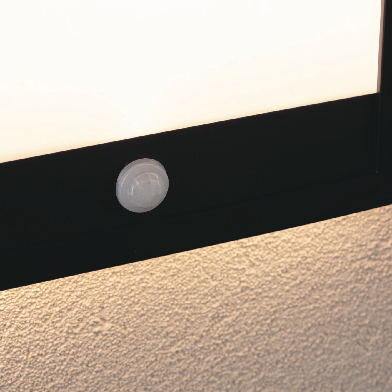Close-up of a Paulmann Outdoor Lamina black frame light, featuring a motion detection sensor and casting a tunable warm white glow against the textured wall.