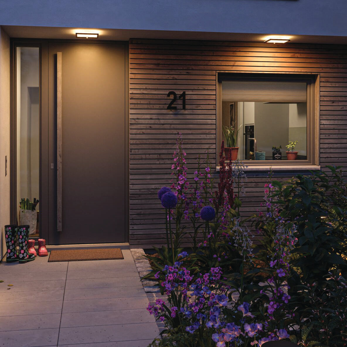 The contemporary home entryway is adorned with wooden paneling, flowers, boots, and vibrant plants by a window at dusk. The Paulmann Outdoor Lamina Insect Friendly 16W LED Backlit Square Tunable Warm Light with Motion Detection in Black illuminates the space with a warm white glow, heightening the inviting atmosphere.