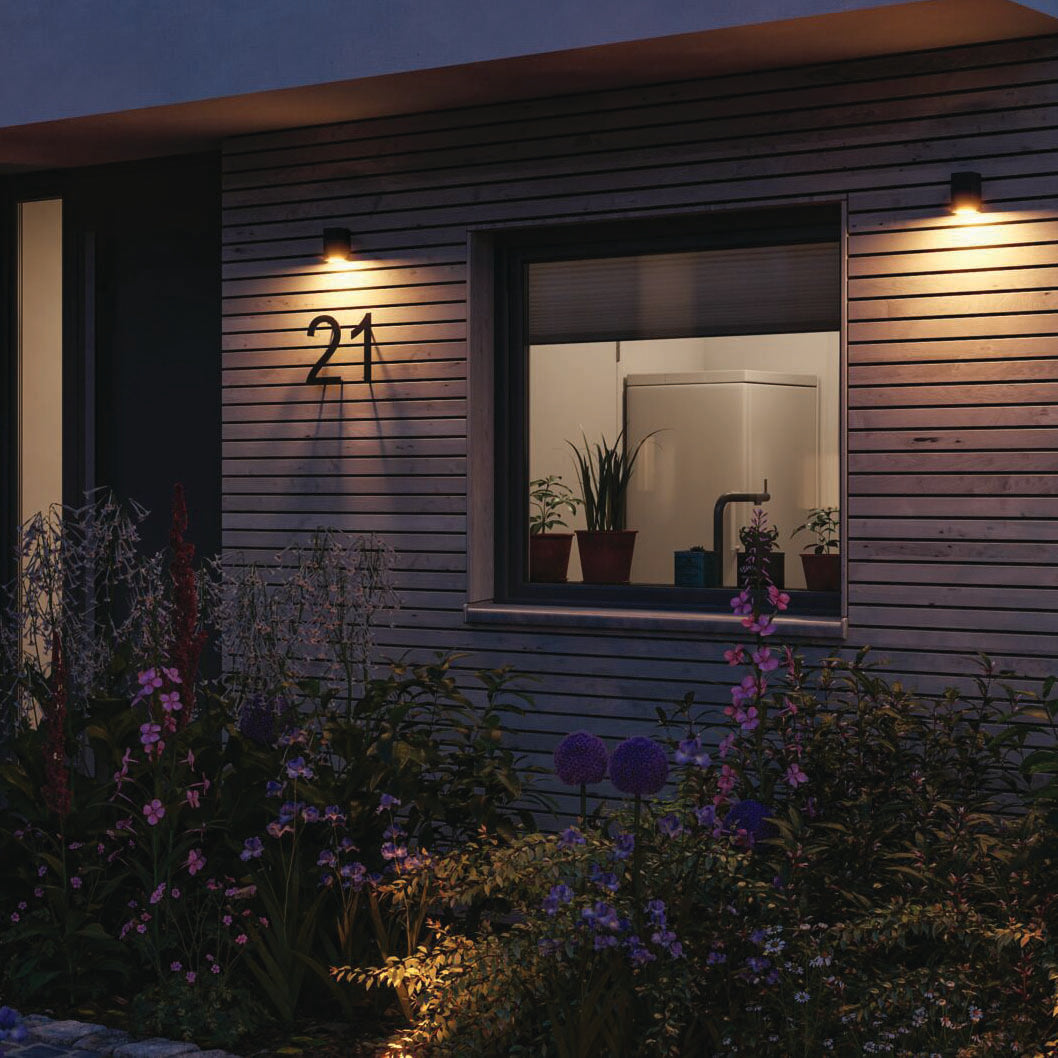 At dusk, the house exterior is adorned with flowers and plants, gently lit by the Paulmann Outdoor Flame Insect Friendly 5.2W LED Single Output Wall Light in Anthracite, with the number 21 displayed beside a window.