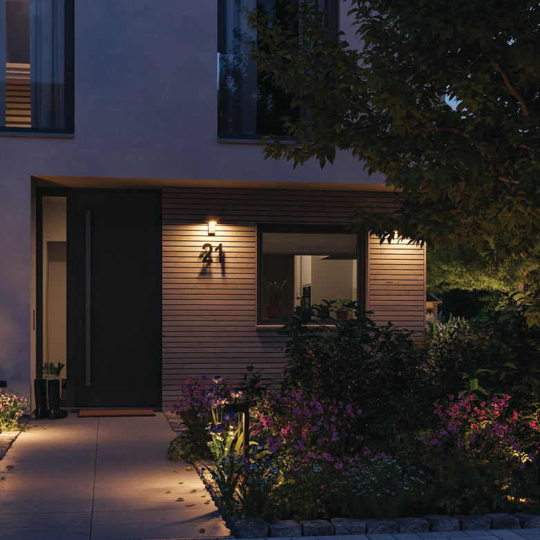 The illuminated modern house entrance, featuring wooden paneling and surrounded by a landscaped garden at dusk, boasts the Paulmann Outdoor Capea 8W LED Wall Light in Anthracite, which offers motion detection and insect-friendly lumination for added convenience.