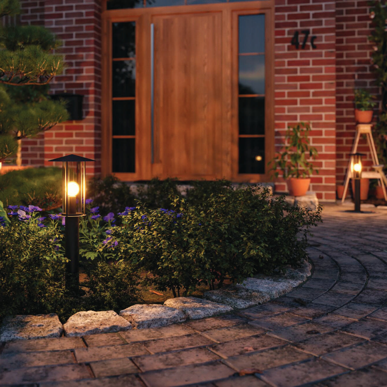 A brick pathway illuminated by Paulmann's Outdoor Classic 15W LED Bollard Lights in Black leads to a wooden door, surrounded by plants and a small ladder adorned with insect-friendly potted blooms.