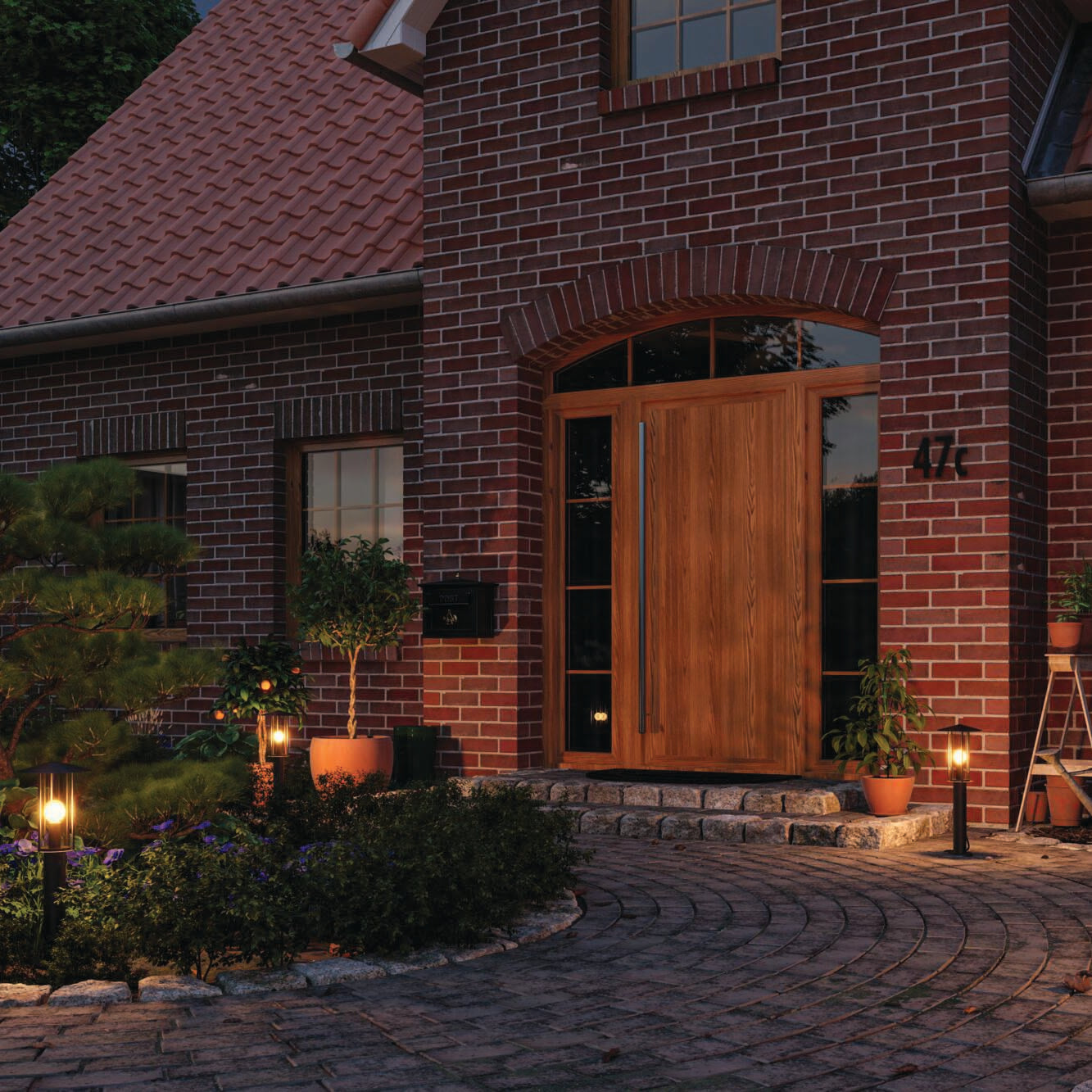 A brick house at dusk with a wooden door, illuminated by Paulmann's Outdoor Classic 15W LED Bollard Lights in Black that are designed to be glare-free. A pathway lined with insect-friendly plants guides you to the entrance.