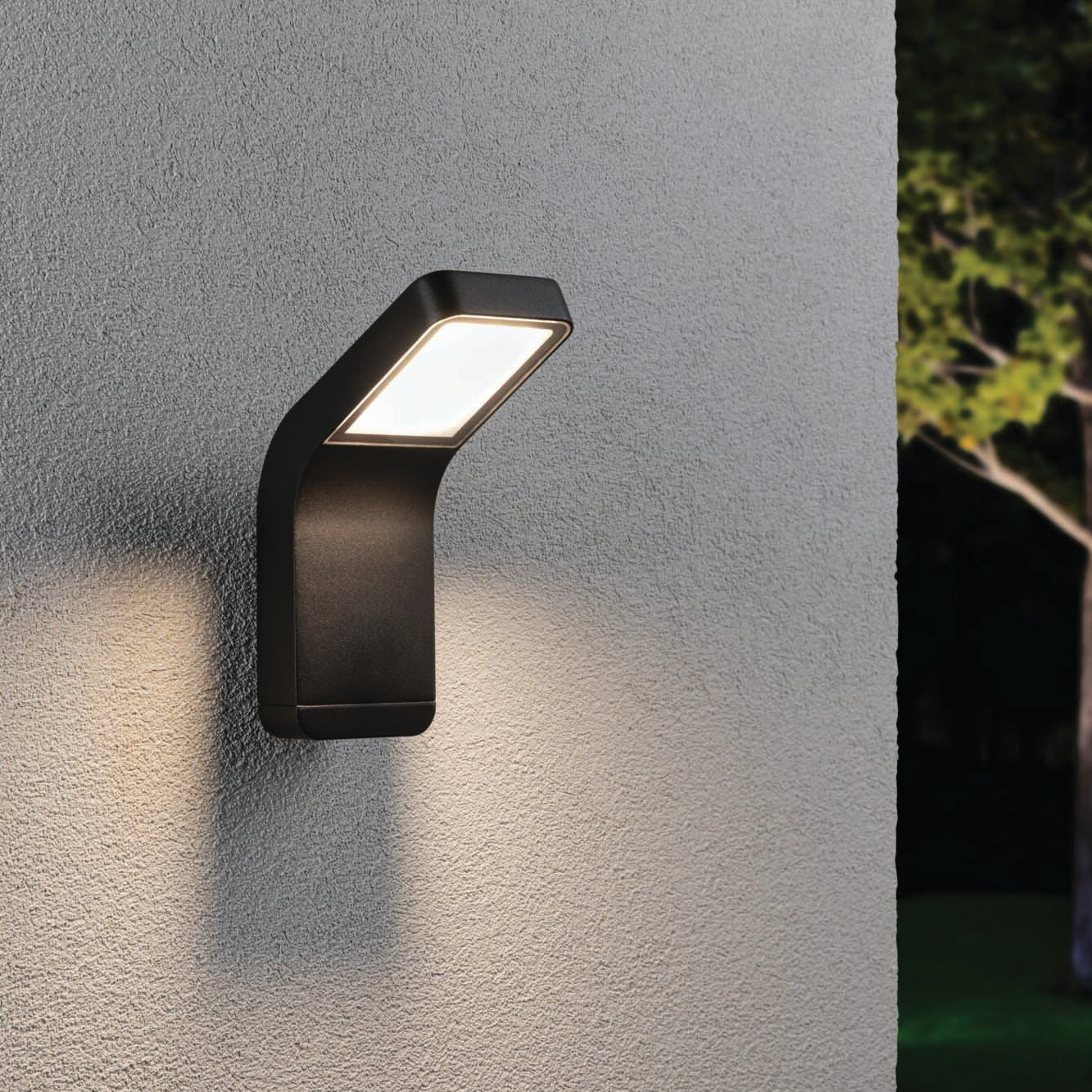 The Paulmann Outdoor Kulma 9W LED Wall Light in Anthracite beautifully lights up a textured wall, with a tree visible in the background. This durable, corrosion-resistant fixture ensures long-lasting elegance and creates a stunning visual impact even in coastal outdoor environments.