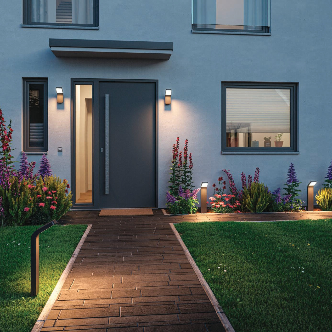 The modern house entrance features a wide door and an illuminated path enhanced by the Paulmann Outdoor Kulma 9W LED Wall Light in Anthracite, with a vibrant flower garden on either side.
