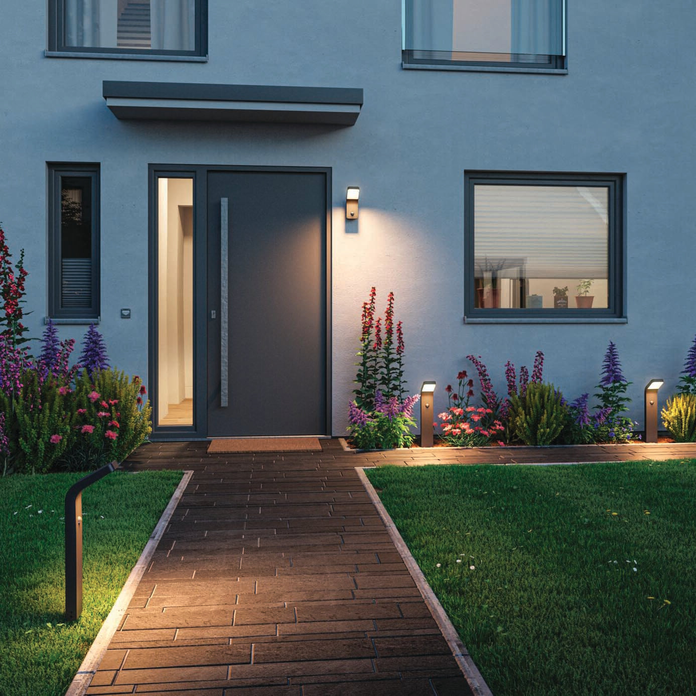 At twilight, a modern house entrance is beautifully illuminated by Paulmann's Outdoor Kulma 9W LED Wall Lights with Motion Detection in Anthracite, guiding visitors along a lit pathway to the large door. The flower garden thrives under their subtle glow, while the corrosion-resistant fixtures ensure durability in any weather.