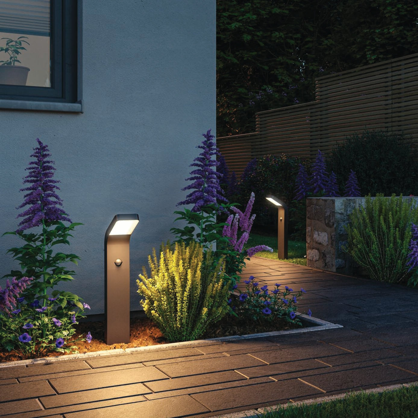 A building's garden pathway illuminated by the Paulmann Outdoor Kulma 9W LED Bollard Light in anthracite with motion detection, surrounded by vibrant purple and green plants.