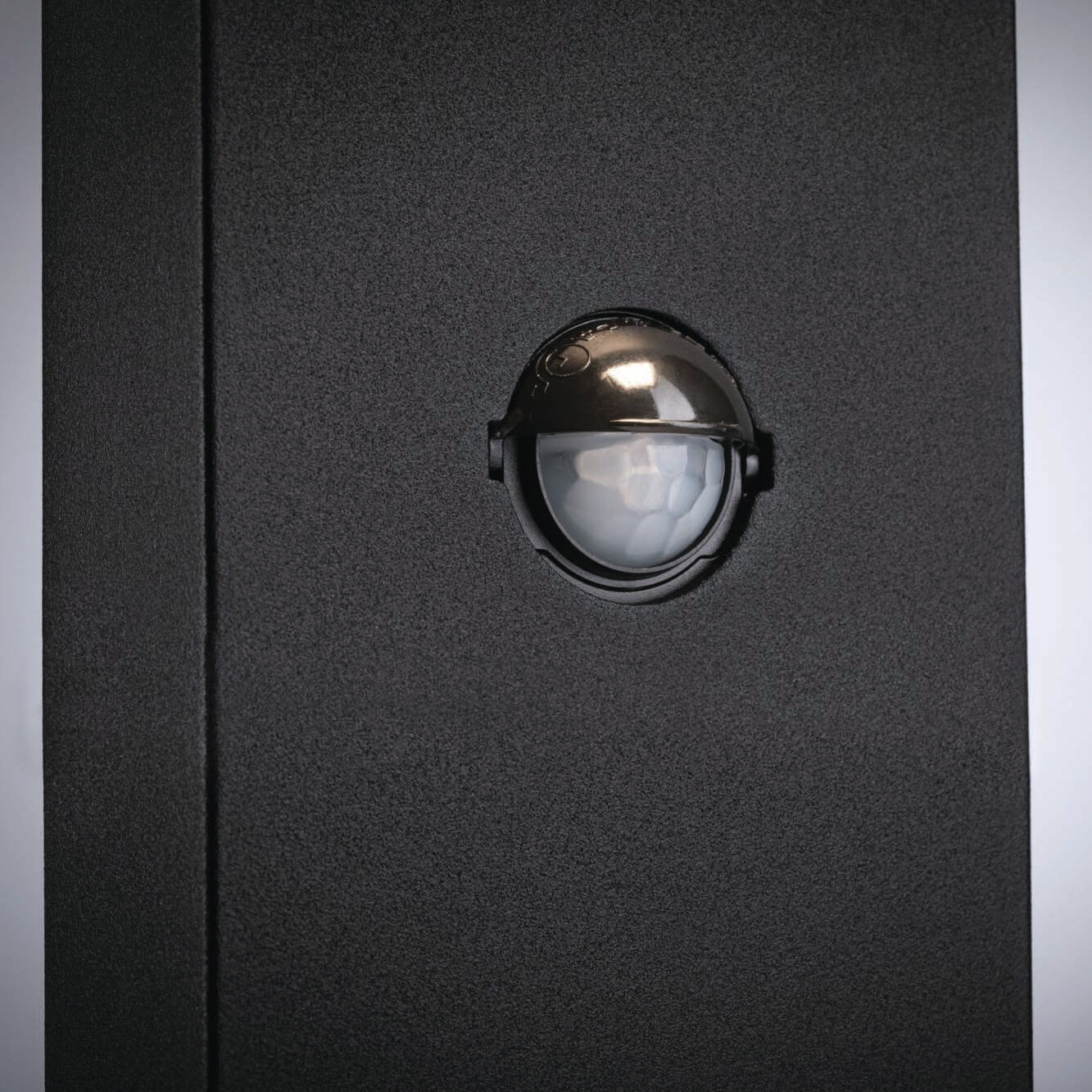 A close-up of a black textured wall showcases a dome-shaped security camera lens at its center, subtly highlighted by the Paulmann Outdoor Kulma 9W LED Bollard Light with Motion Detection in Anthracite for enhanced visibility.