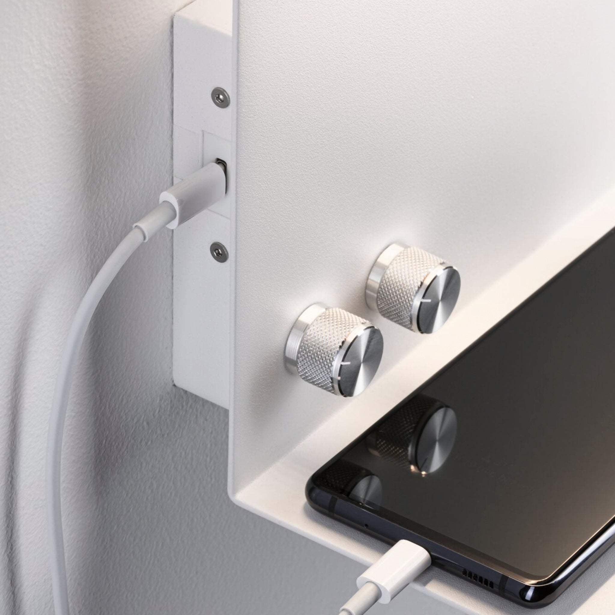 A smartphone charges on a wall shelf with silver knobs, using a sleek white USB-C cable, positioned directly below the Paulmann - Jarina 7.5W LED Dimmable Wall Light with USB Port in White Matt.