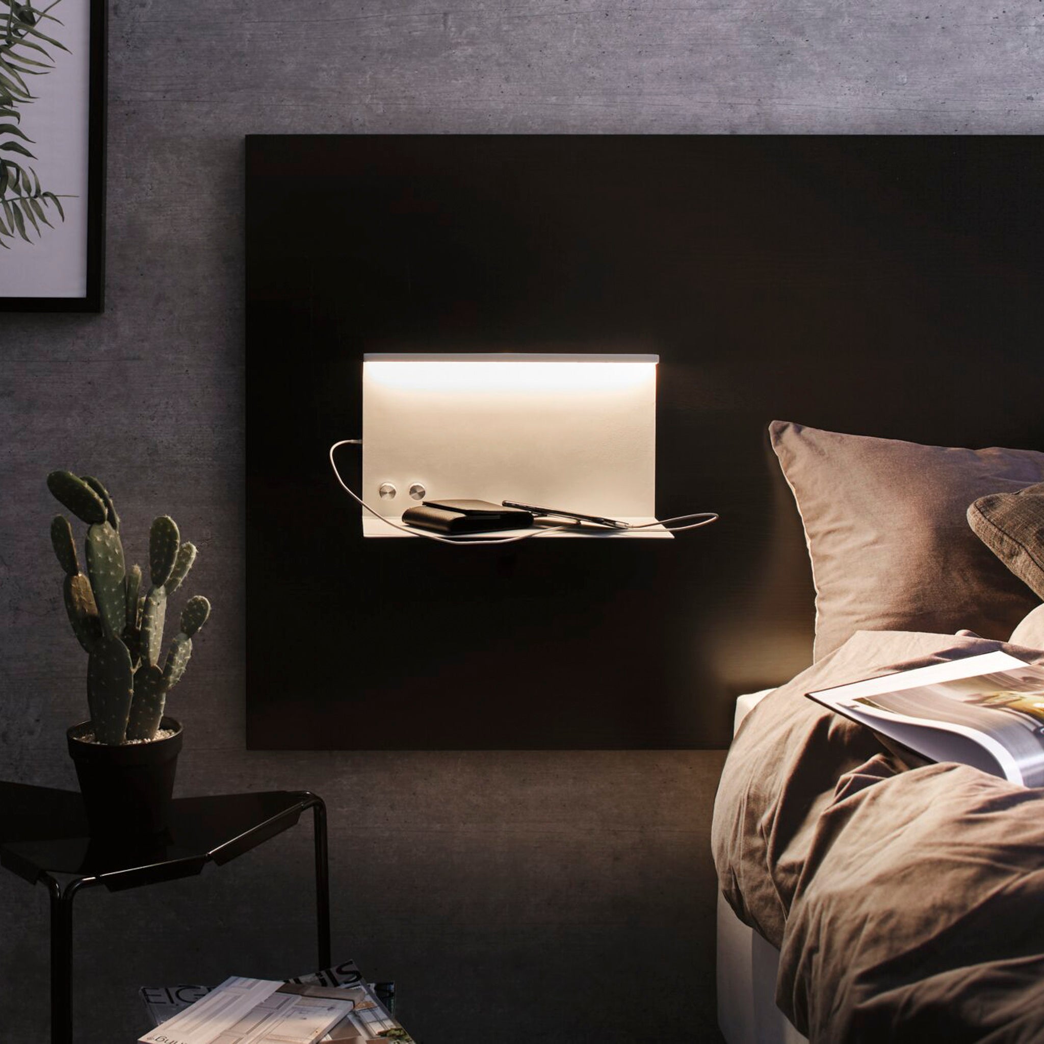 A bedside setting includes the Paulmann Jarina 7.5W LED dimmable wall light in white matt, which features a modern lighted shelf with a sleek USB-C charging port, perfect for accommodating a smartphone and magazine. This wall-mounted luminaire casts a warm and inviting glow, while a potted cactus on the table introduces an element of nature.