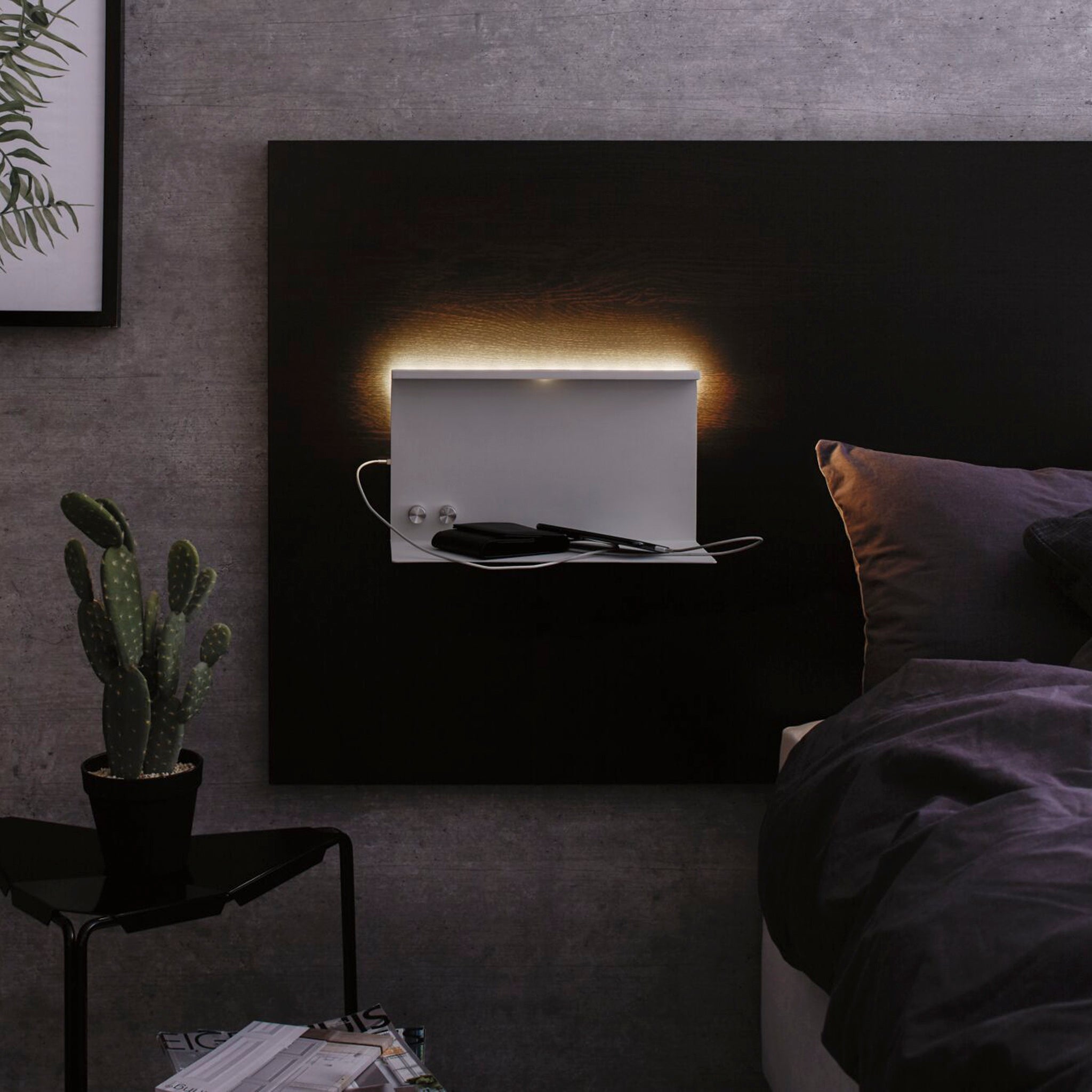 In a modern bedroom, there's an illuminated wall shelf and a cactus adorning the side table beside the bed. The minimalist decor, highlighted by dark tones, features the sleek and functional Paulmann Jarina 7.5W LED Dimmable Wall Light with USB Port in White Matt for wall-mounted reading.