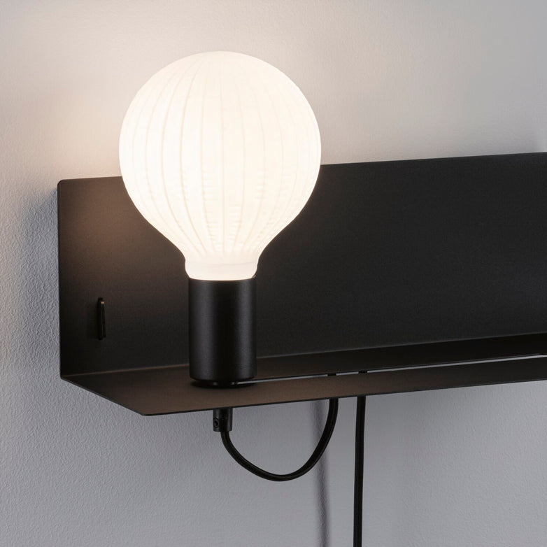 The Paulmann - Devara 40W Wall Mounted Lamp with USB Port in Black Matt showcases a modern design featuring a large frosted bulb and rectangular backing, complemented by an integrated shelf and USB-C charging port, all set against a white wall.