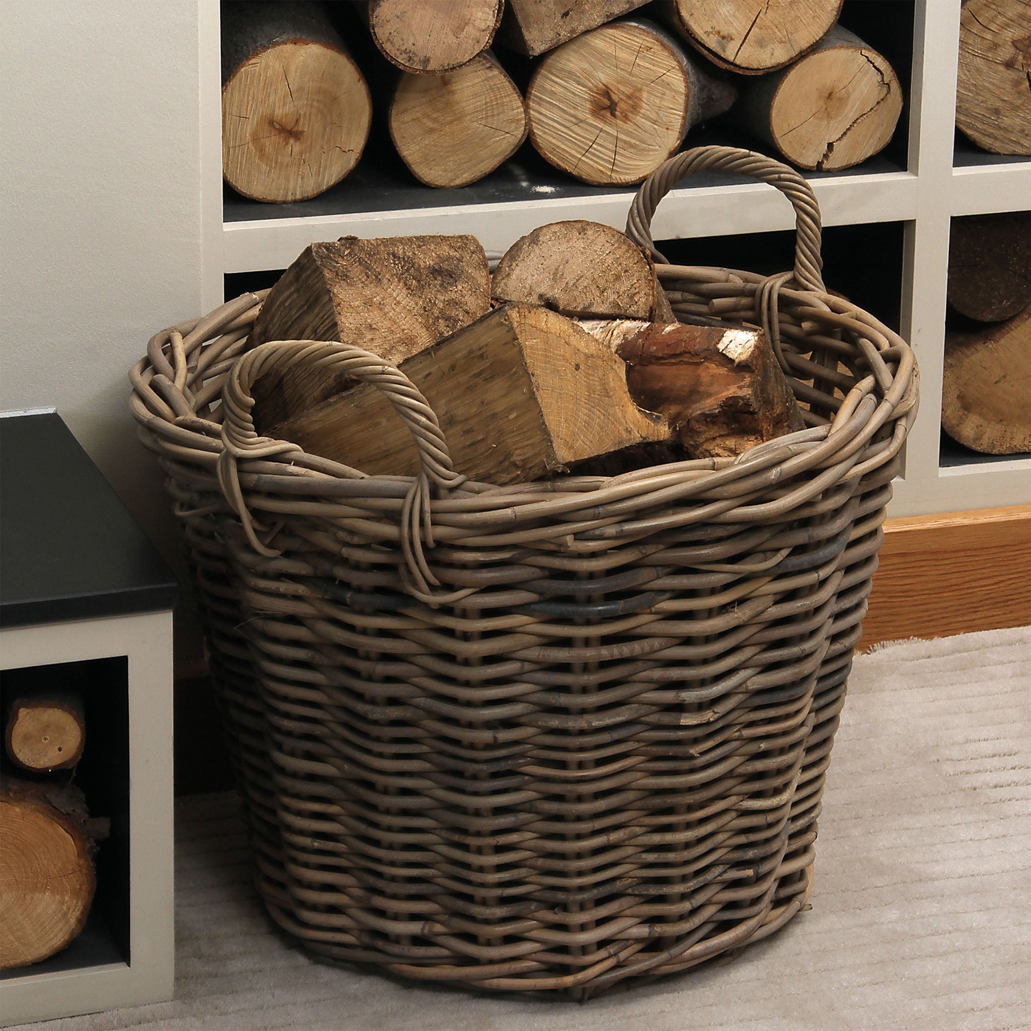 A Pacific Lifestyle - Bali Grey Kubu Round Storage Basket filled with firewood rests on a beige carpet, beautifully enhancing the nearby wood storage shelf.