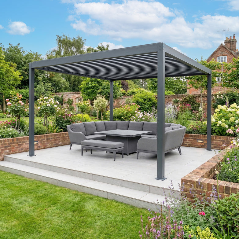 The PergoSTET Plus Frame Only 4m x 4m Square Pergola with LED Lighting in Grey sits on the patio, paired with a sectional sofa and table, and surrounded by a vibrant, colorful garden.