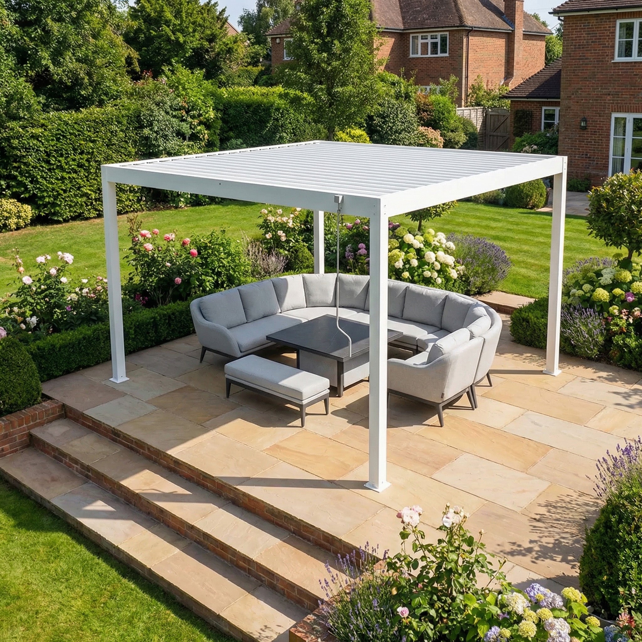 A PergoSTET Plus Frame Only 4m x 4m Square Pergola with LED Lighting in White shelters a gray outdoor sectional on a stone patio in a lush garden.