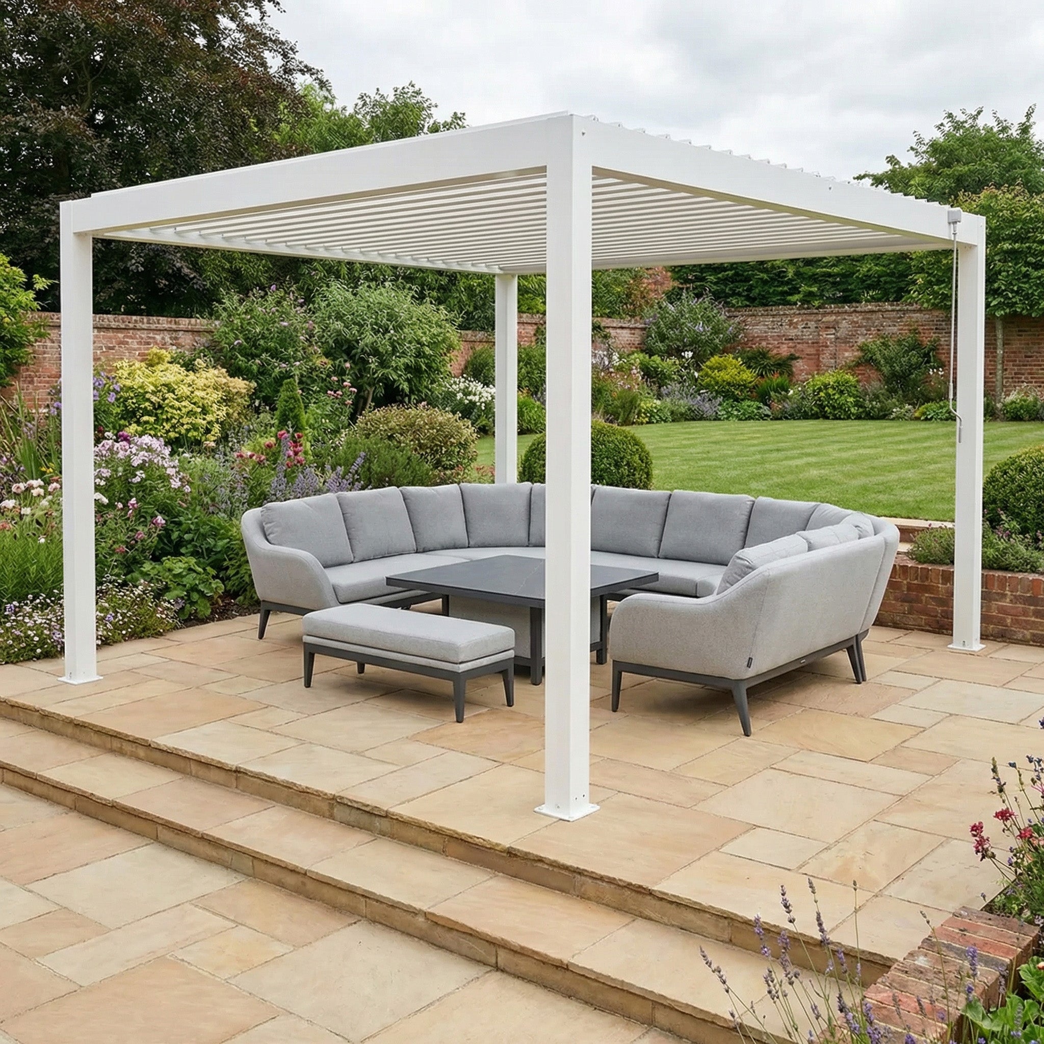 Modern patio with the PergoSTET Plus 4m x 4m Square Pergola Frame in White featuring LED lighting, paired with a grey sectional sofa and table on a stone terrace in a lush garden.