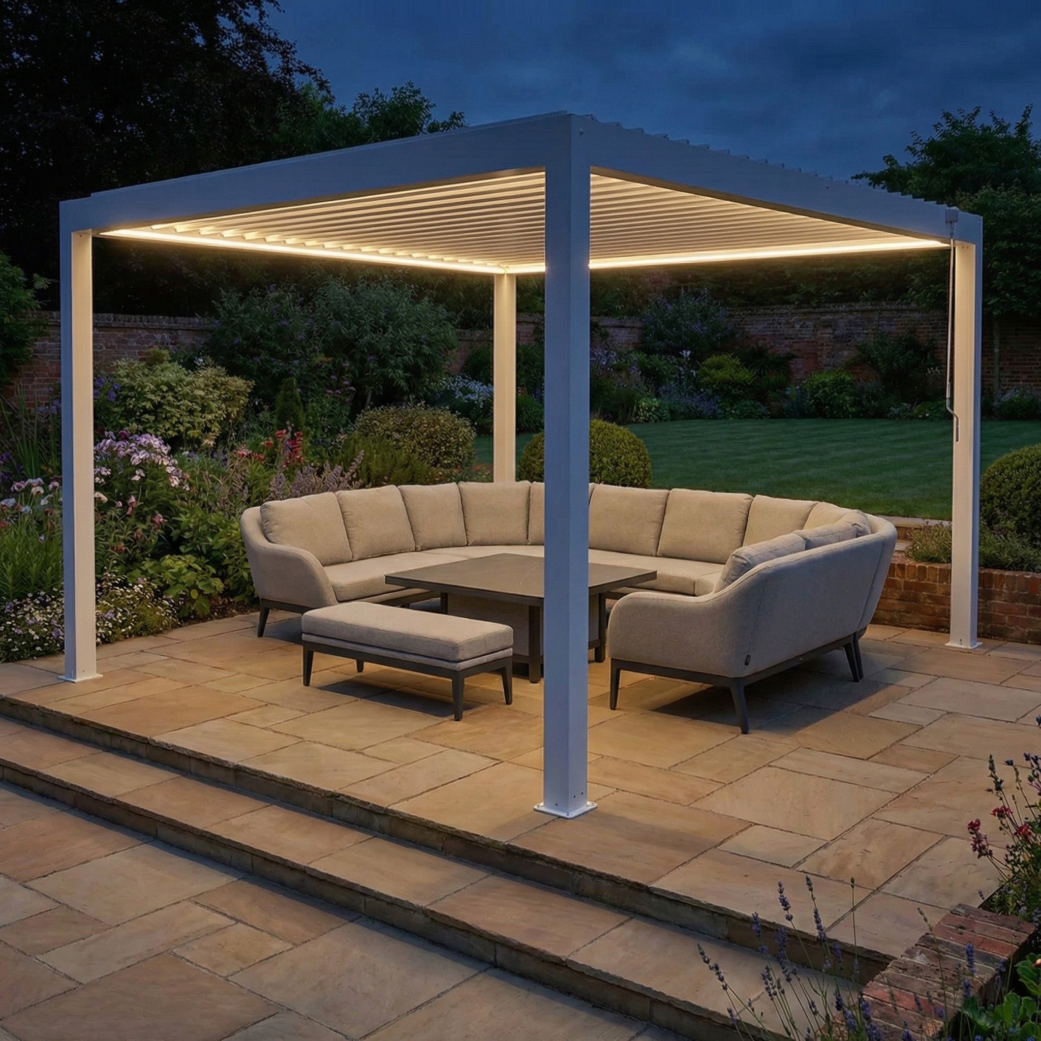 A modern outdoor patio features the PergoSTET Plus Frame Only 4m x 4m Square Pergola with LED Lighting in White, paired with a sectional sofa, ottoman, and central table on stone tiles at dusk.