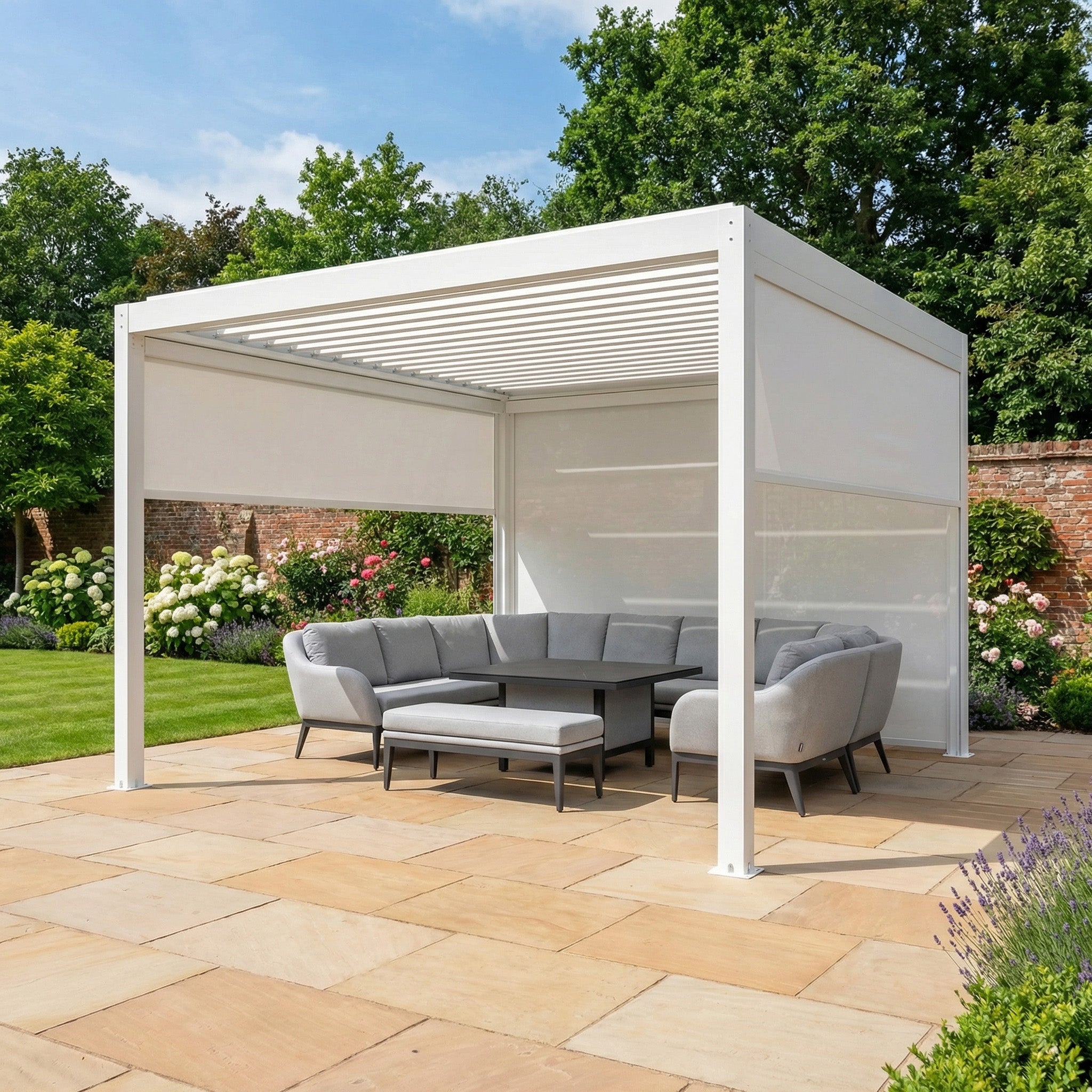The PergoSTET Plus 4m x 4m Square Pergola in white, featuring a louvred roof, LED lighting, and three manual side blinds, stands on a stone terrace with gray patio furniture in a landscaped garden.