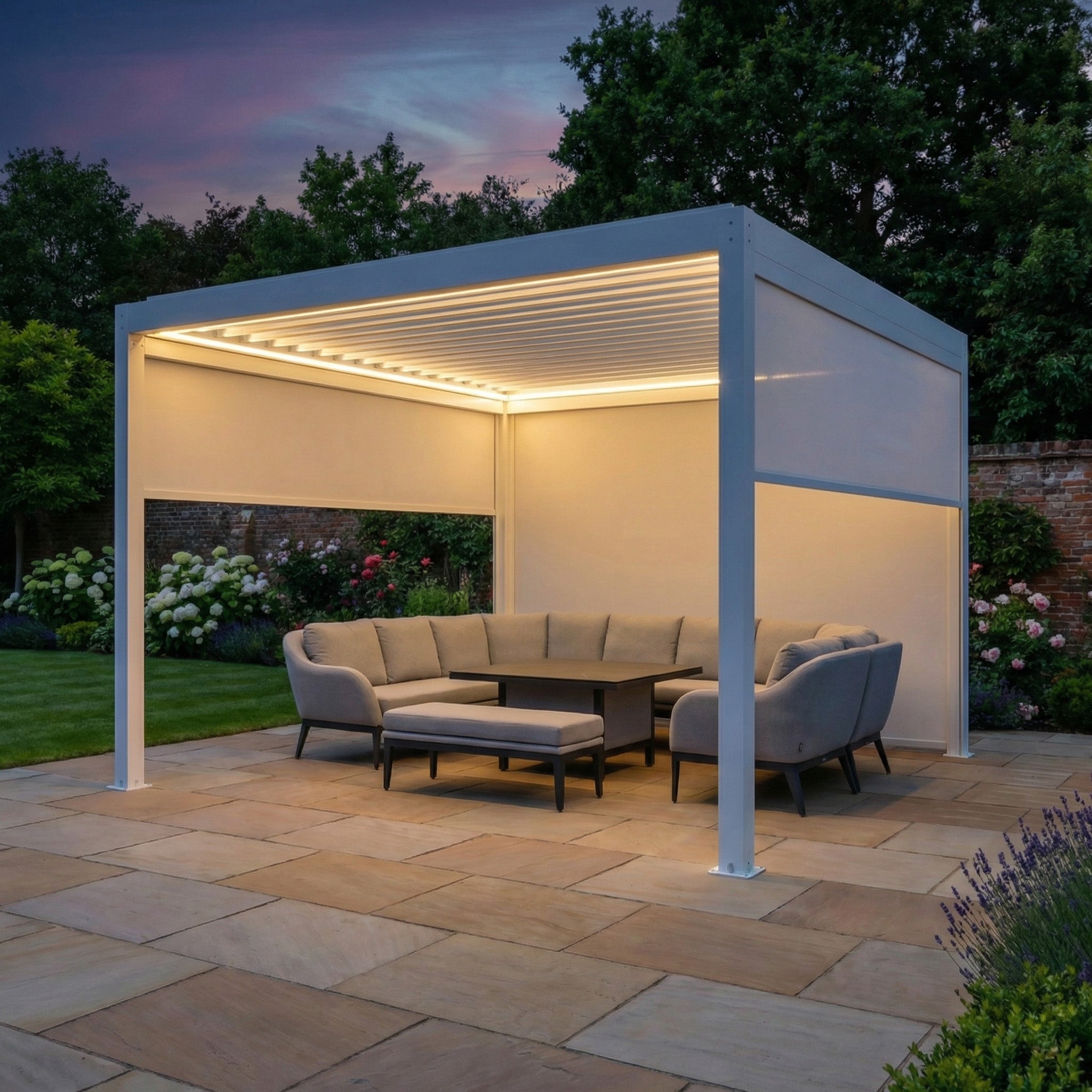 The PergoSTET Plus 4m x 4m Square Pergola in white features LED lighting and 3 manual side blinds, shown at dusk with a gray sectional sofa and chairs on a stone patio.