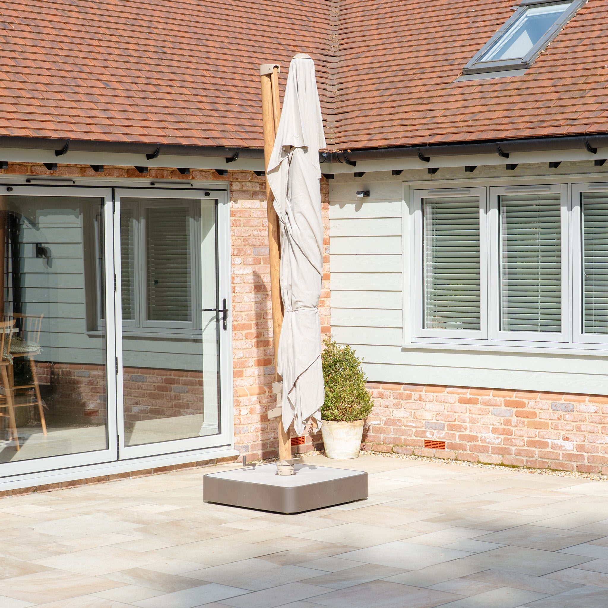 The Finca 4m x 3m Cantilever Parasol in Sand provides sun protection on a sunny patio with a brick and wooden house as a backdrop, creating an inviting outdoor living space.